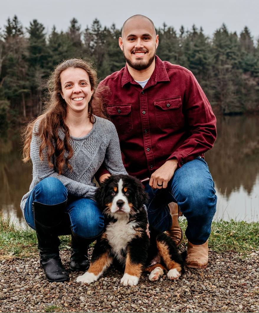Smiling couple crouches beside a Bernese Mountain Dog on a gravel path by a calm lake with evergreen trees in the background.