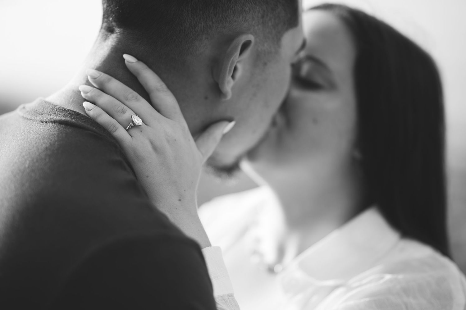 Black and white photo of a couple sharing a romantic kiss as the woman's hand with a sparkling engagement ring rests on his neck.