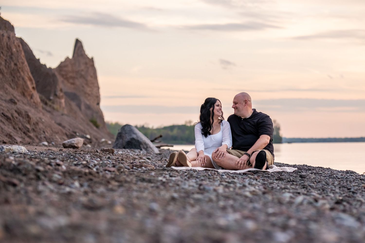 Couple sitting close together on a blanket on a rocky shoreline, smiling at sunset with cliffs and calm water behind them.