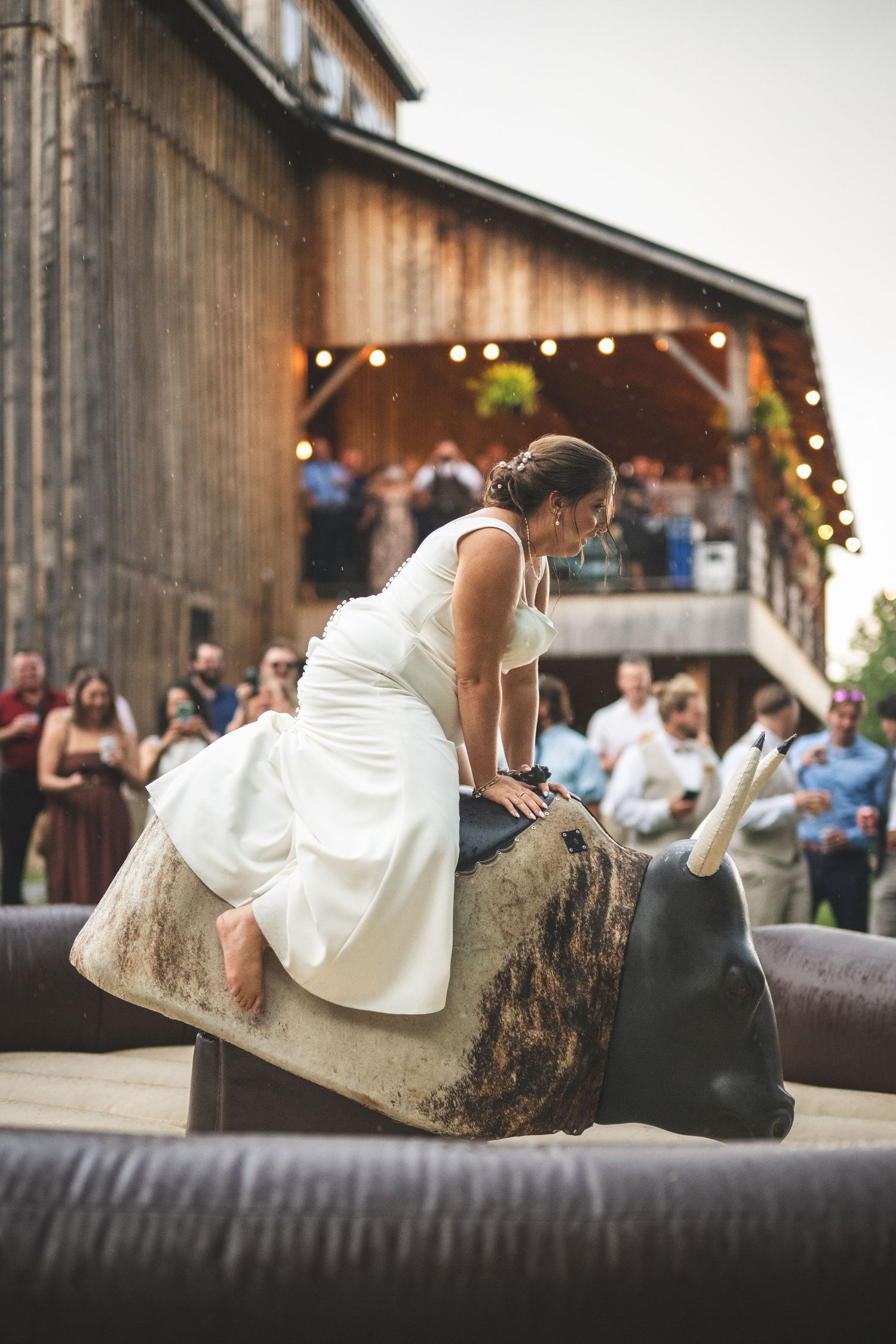 Bride in a white wedding dress rides a mechanical bull at a rustic barn reception while guests cheer and watch from nearby.