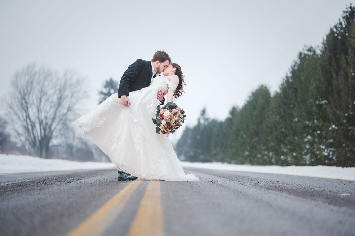 Bride and groom share a dipped kiss on a snowy country road, her white gown flowing and holding a bouquet, with evergreen trees behind.