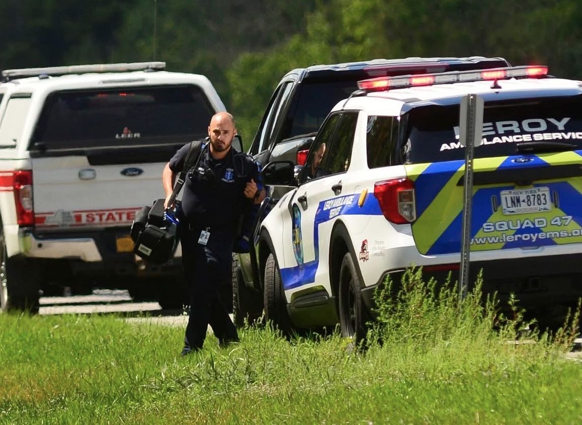 Paramedic in dark uniform holding a helmet walks across grass beside an ambulance and a pickup truck on a roadside.
