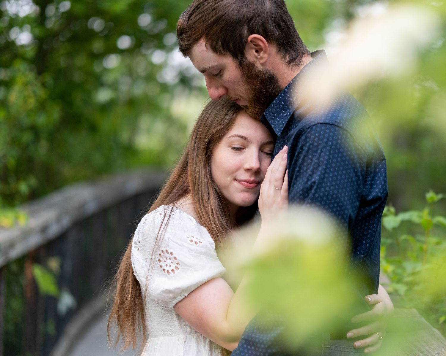 A romantic couple embraces tenderly on a bridge surrounded by soft green bokeh and natural light.
