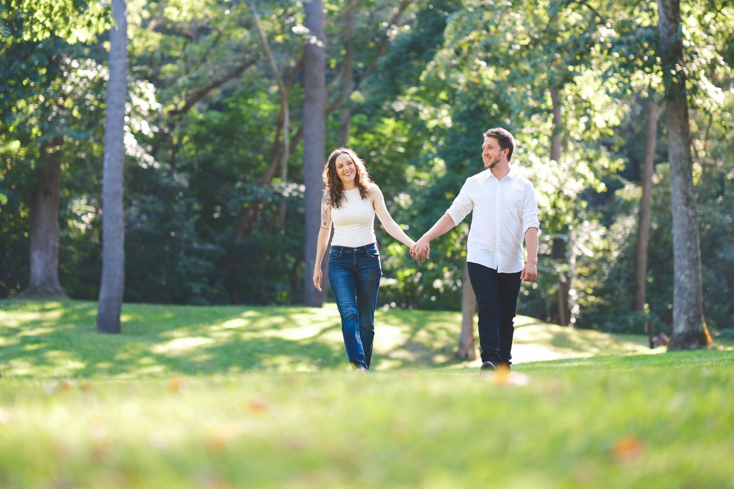A couple holds hands and walks across a sunny grassy park lined with tall trees during an outdoor engagement photo shoot.