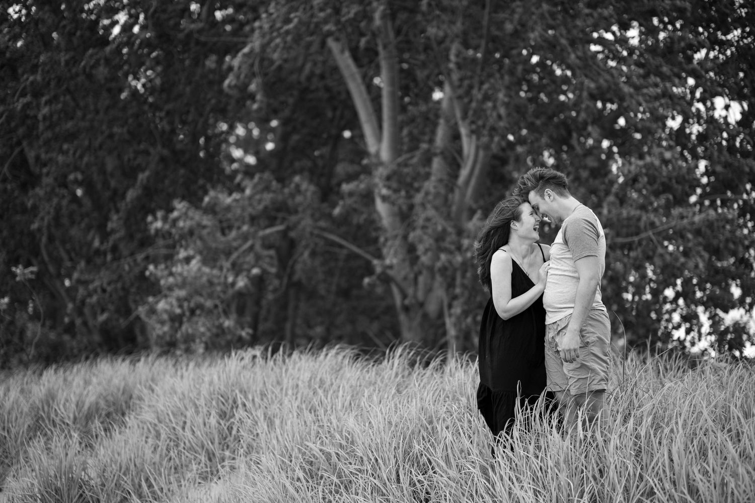 Black and white photo of a couple sharing a close moment in tall grass in front of leafy trees.