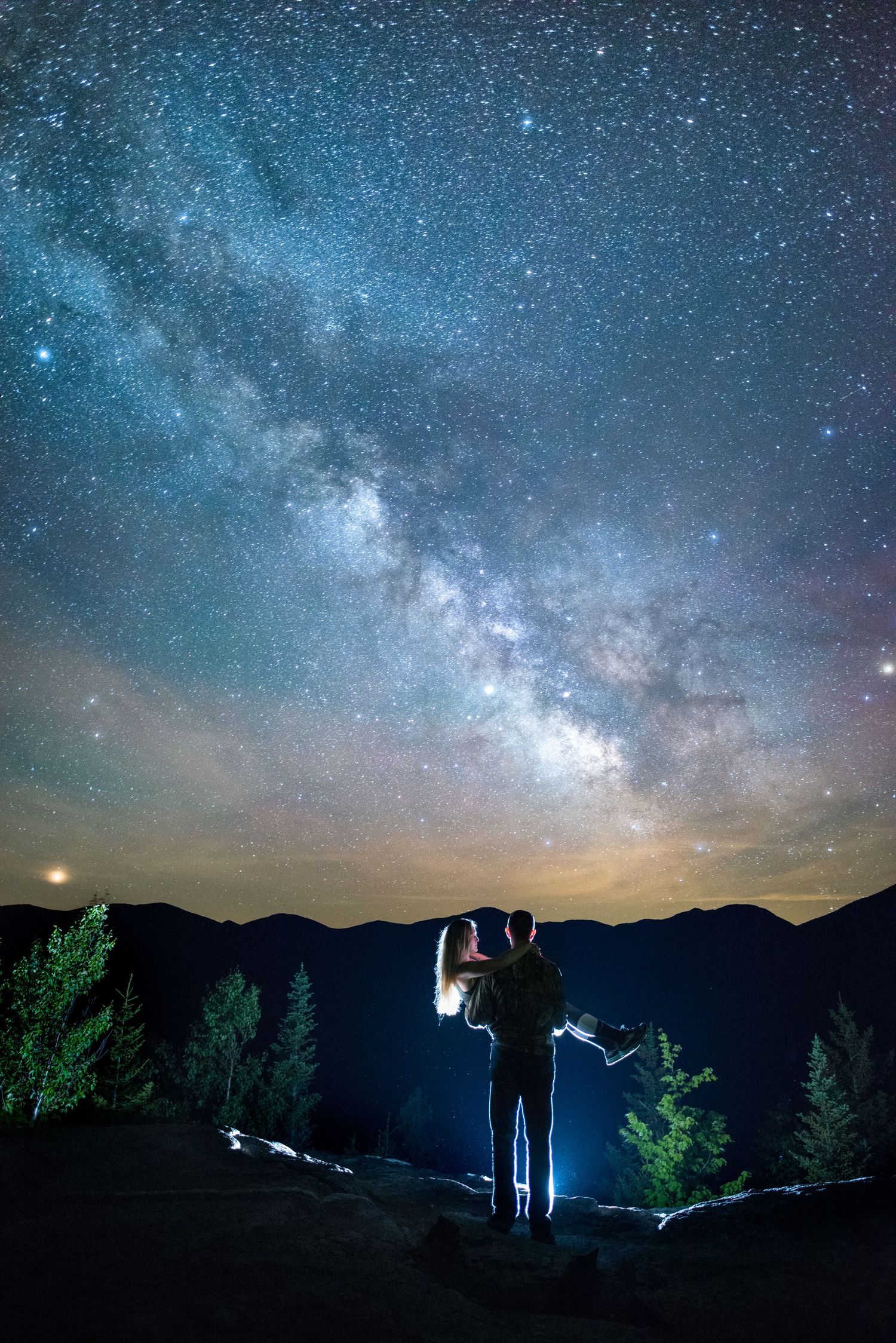 Couple silhouetted on a mountain ridge, embracing under a vivid Milky Way and starry night sky, with pine trees and distant peaks nearby.