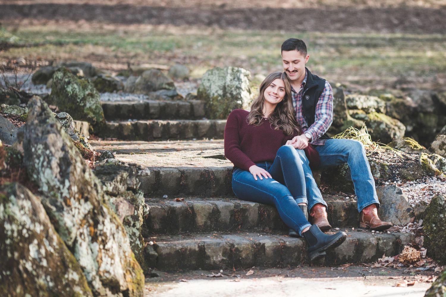 Couple in cozy fall outfits sitting together on stone steps surrounded by mossy rocks for an engagement session.