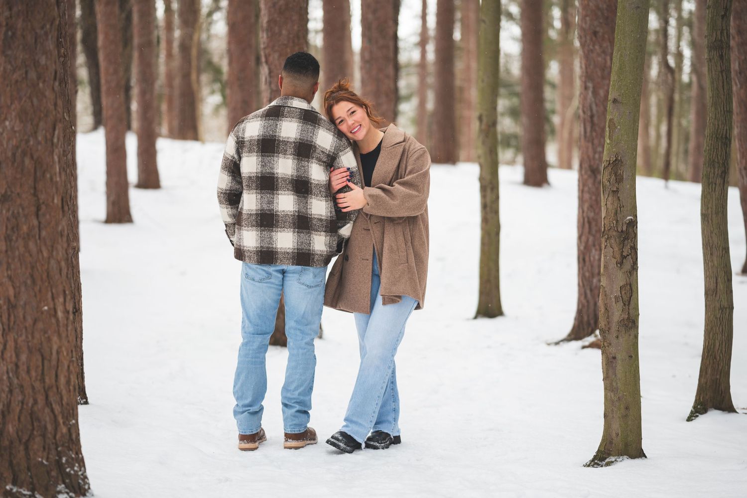 Smiling couple embracing in a snowy pine forest during a winter engagement photo session.