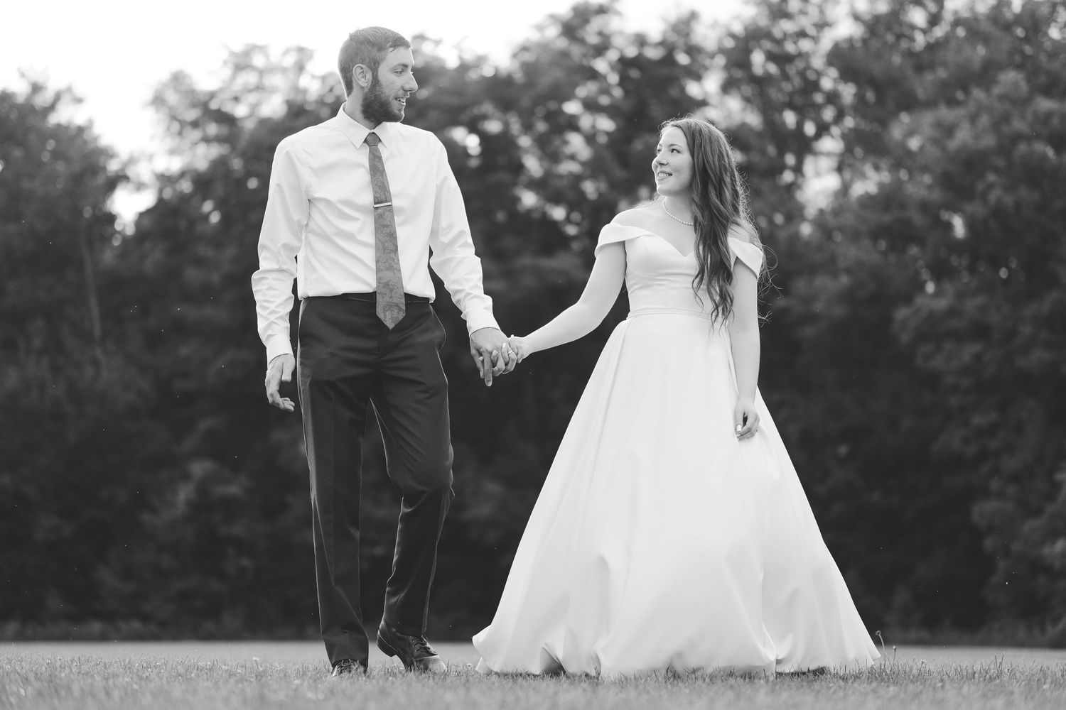Black and white photo of a bride in an off-the-shoulder gown holding hands with a groom in a tie while walking across a grassy field.