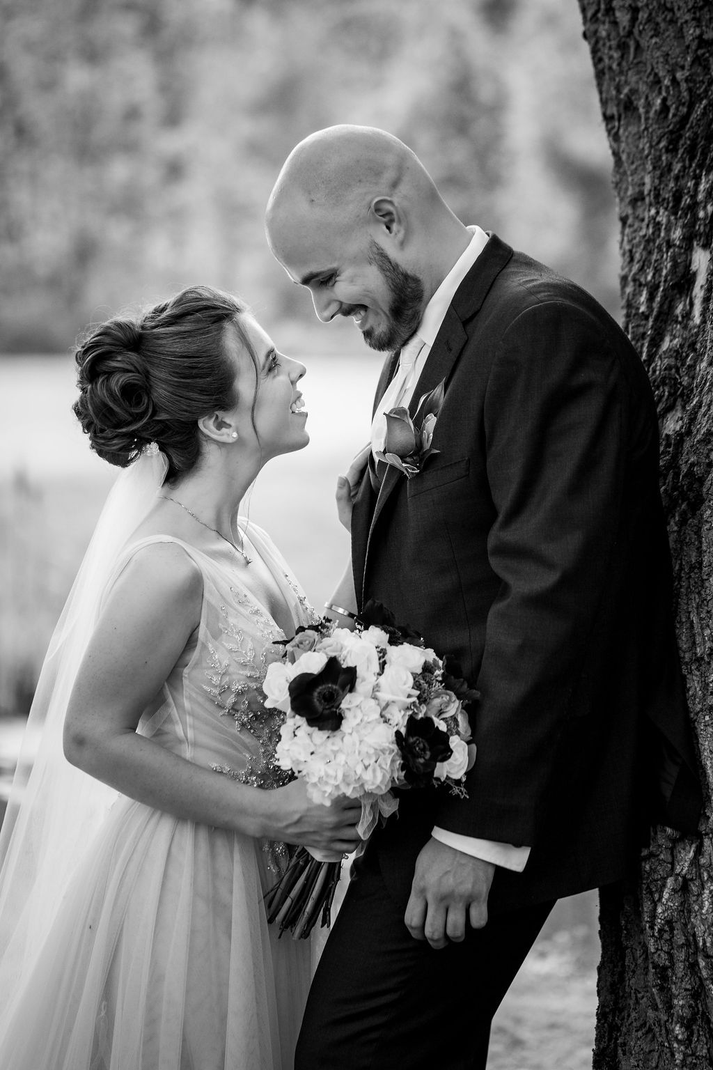 Black and white wedding portrait of a smiling bride holding a bouquet and her groom in a suit leaning against a tree.
