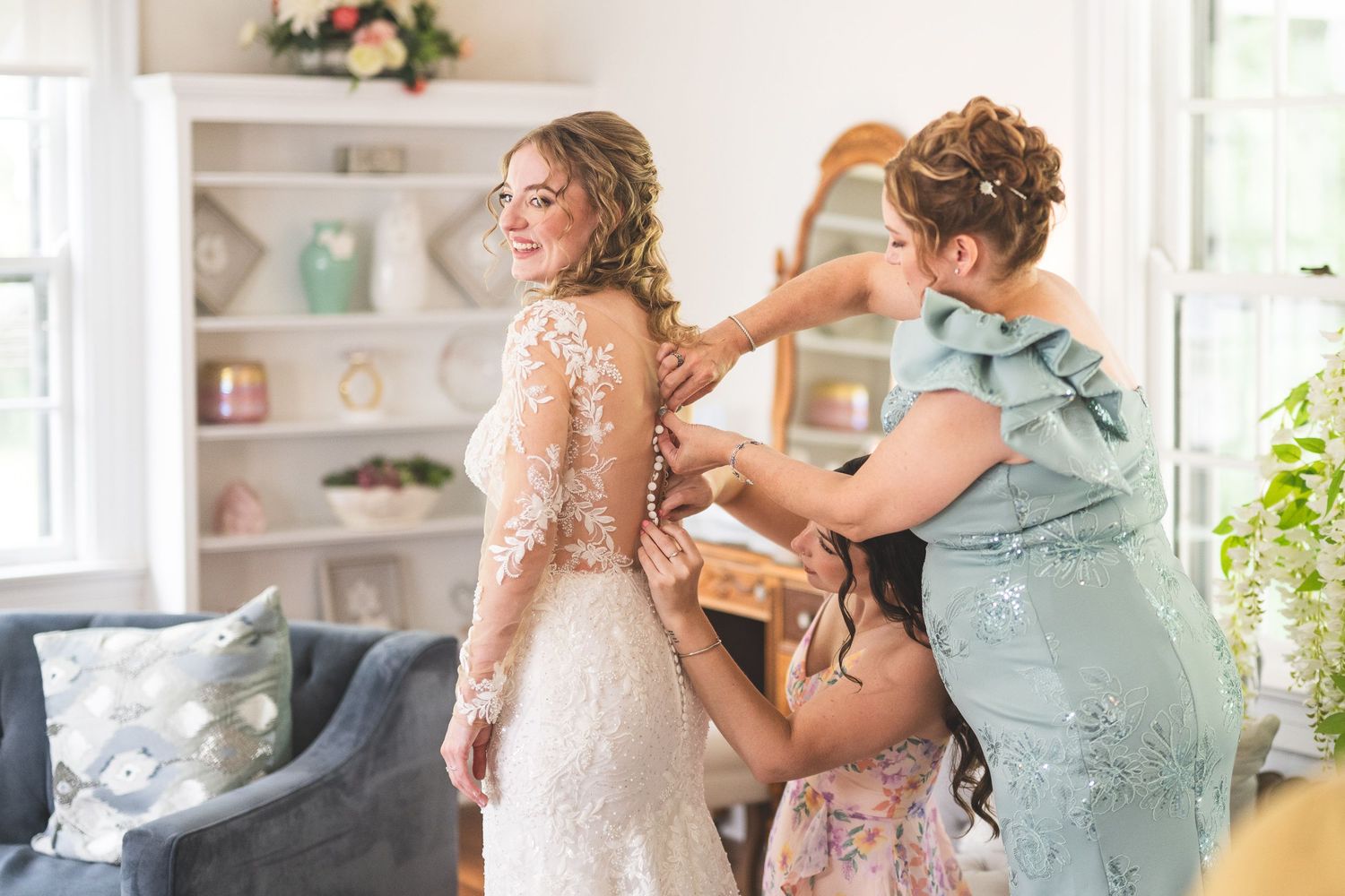 Bride in a lace wedding gown gets help with her dress from two women in a bright room with a gray armchair and white shelves.