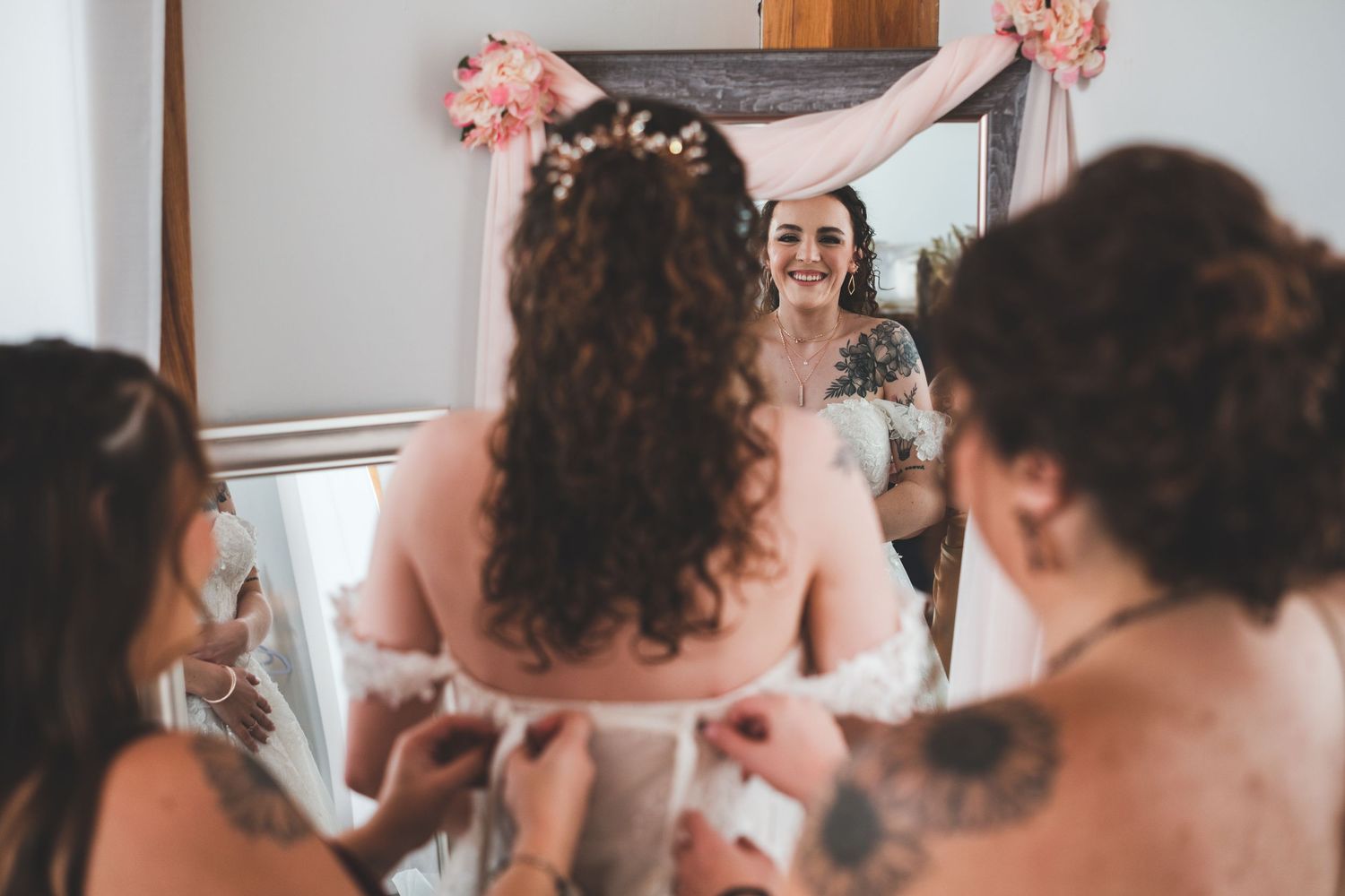 Bride with curled hair and a floral hairpiece faces a mirror as bridesmaids lace up her wedding dress while she smiles in the reflection.