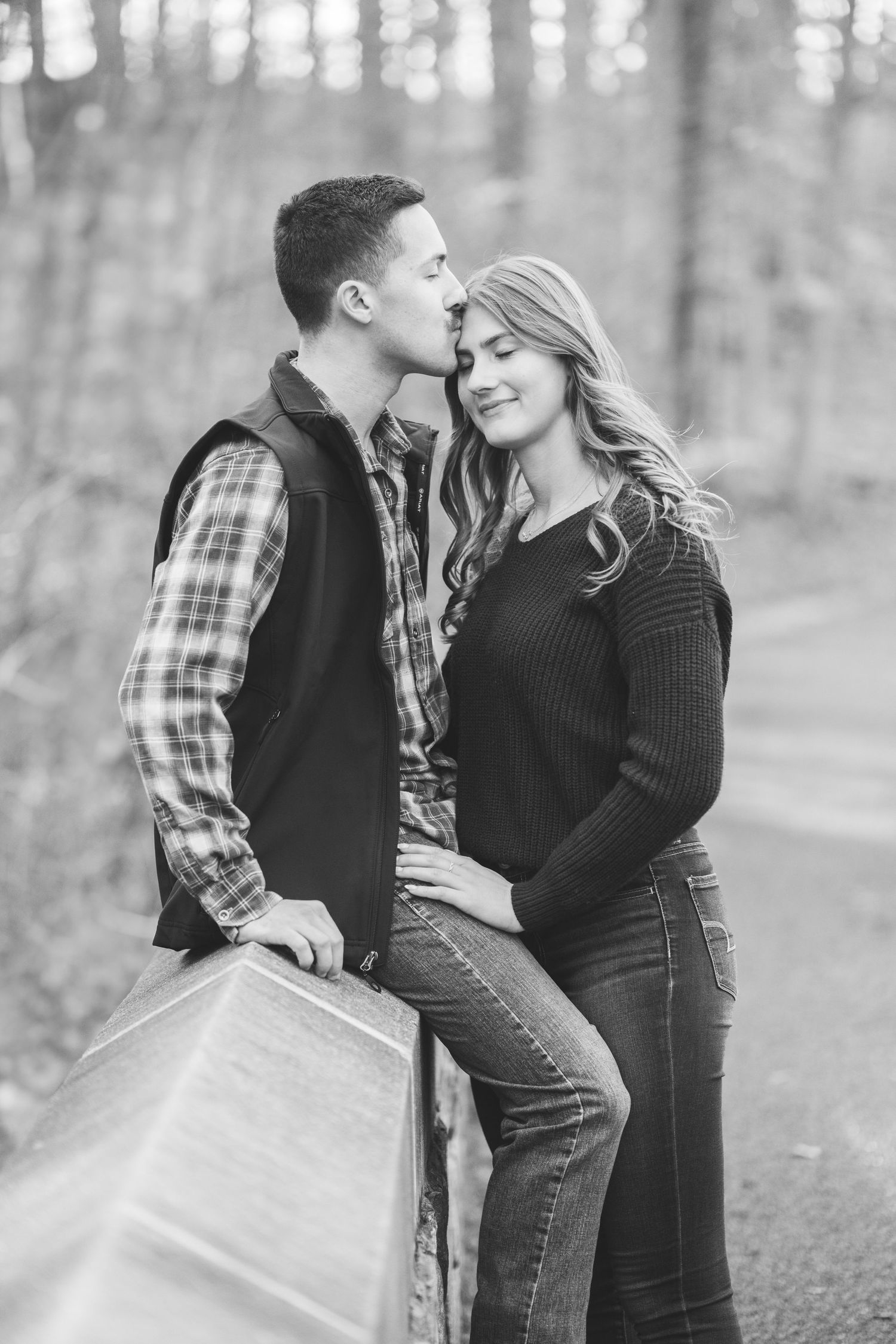 Black and white engagement photo of a man in a plaid shirt and vest kissing a smiling woman's forehead beside a wooden railing in the woods.