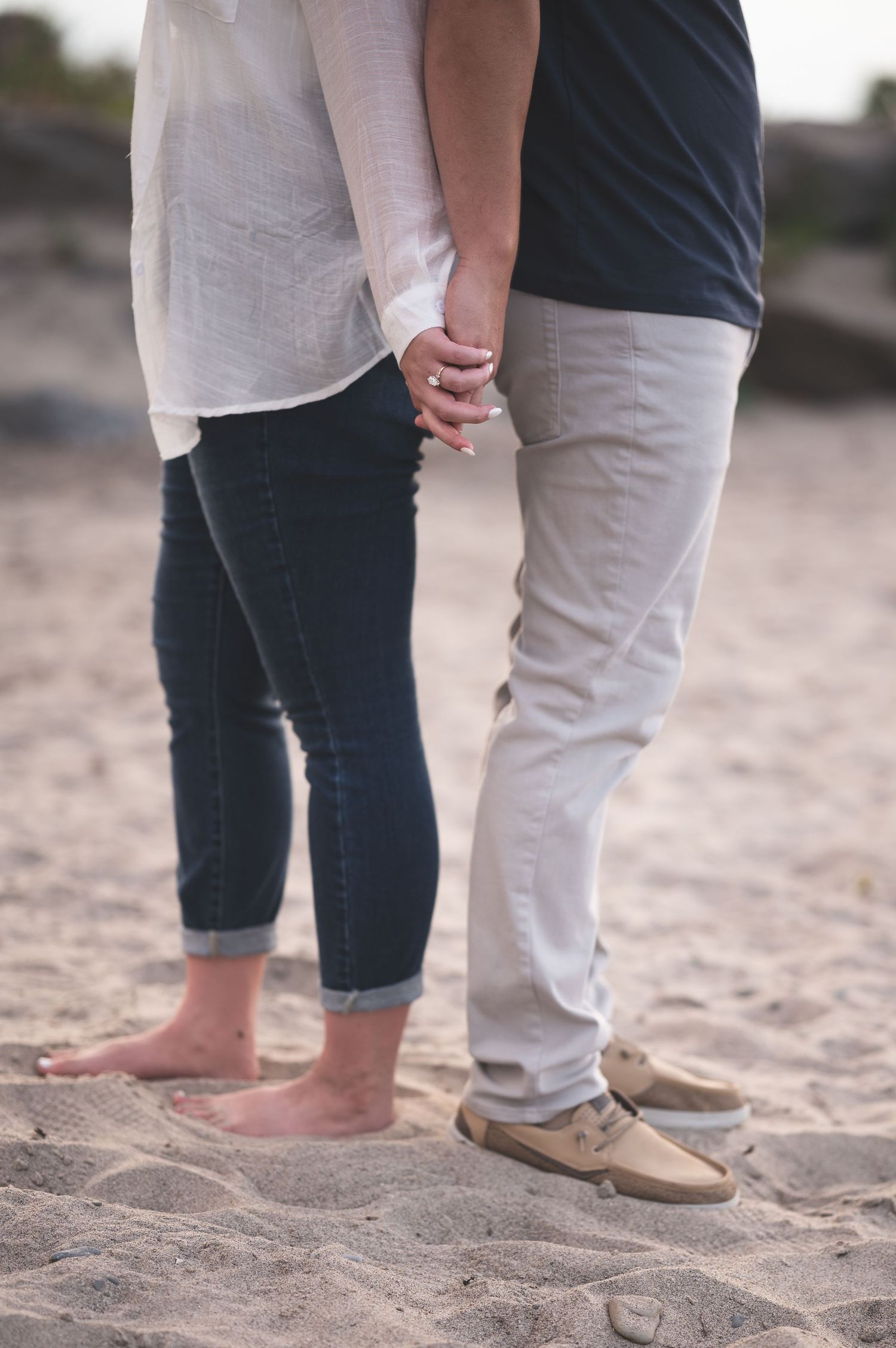 Couple holding hands on a sandy beach, shown from the knees down, with her barefoot in rolled jeans and him in khakis and boat shoes.