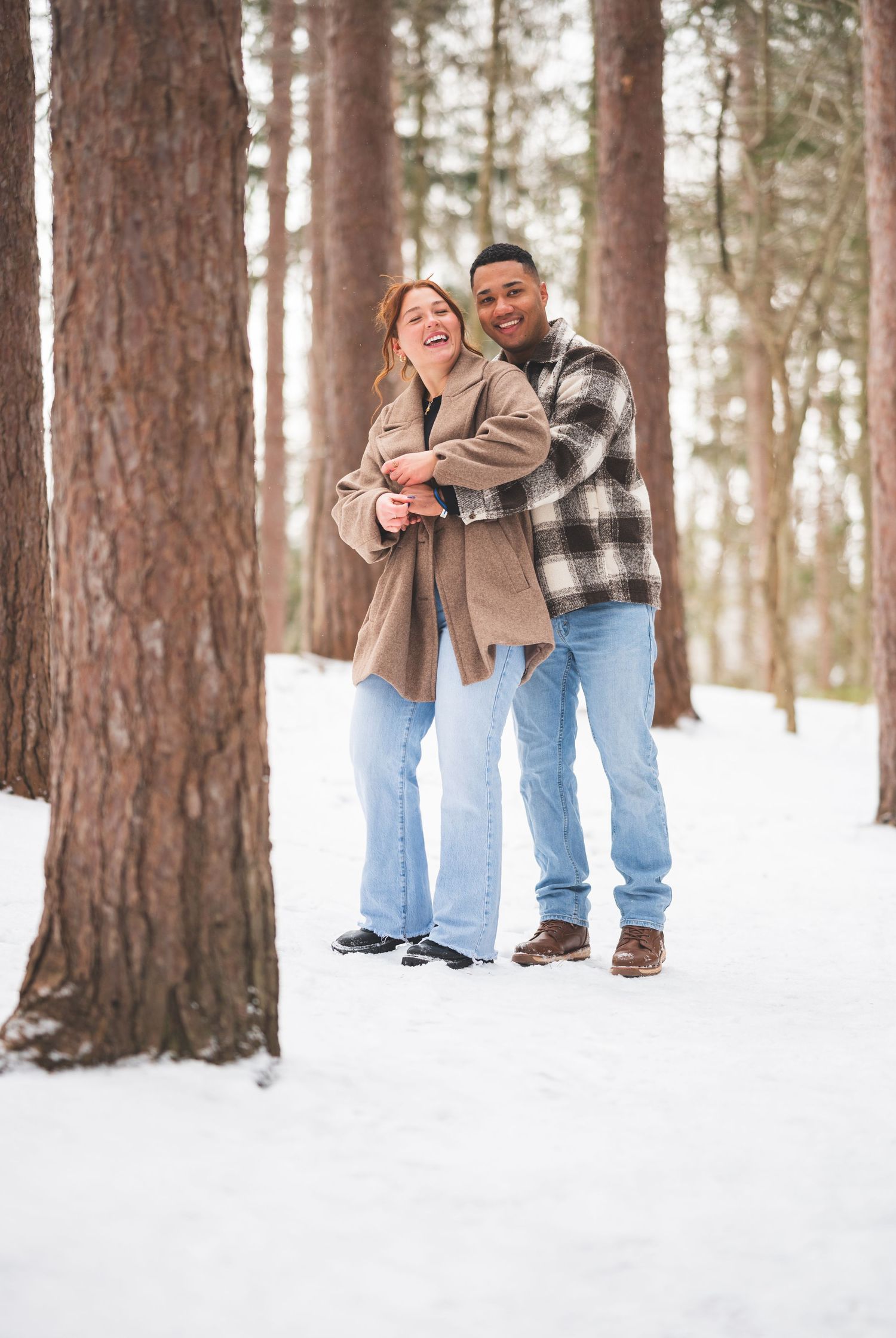 Smiling couple in jeans and cozy coats embrace and laugh among tall pine trees on a snowy winter forest path.