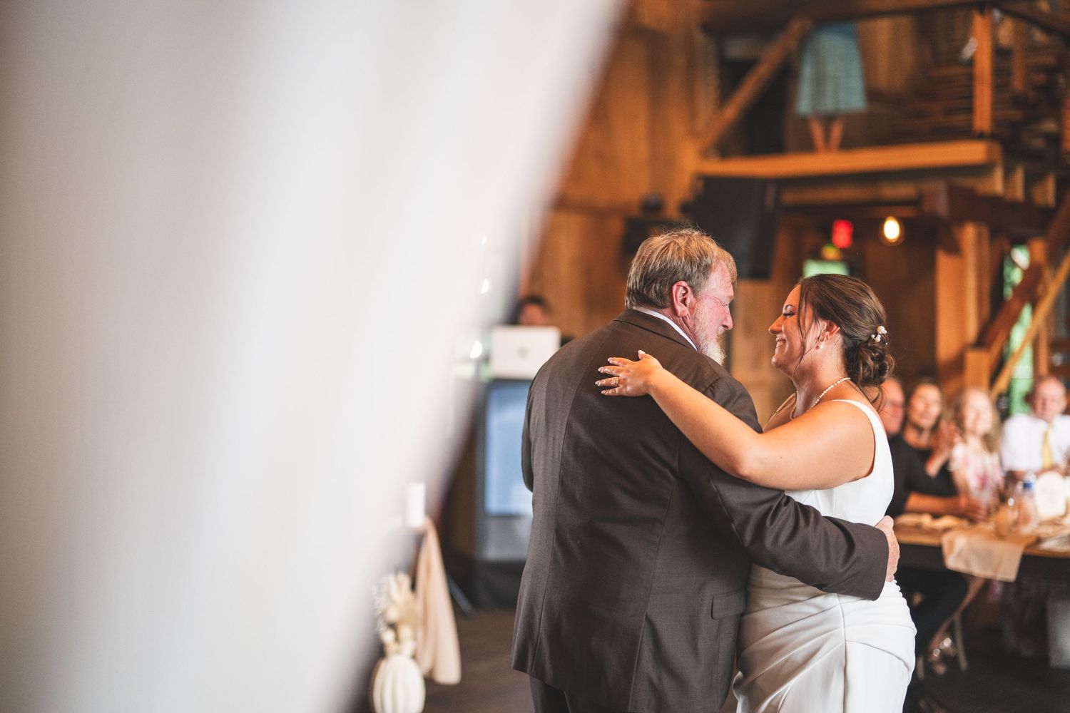 Father and bride in a white gown share an emotional dance at a rustic barn wedding reception while guests watch in the background.