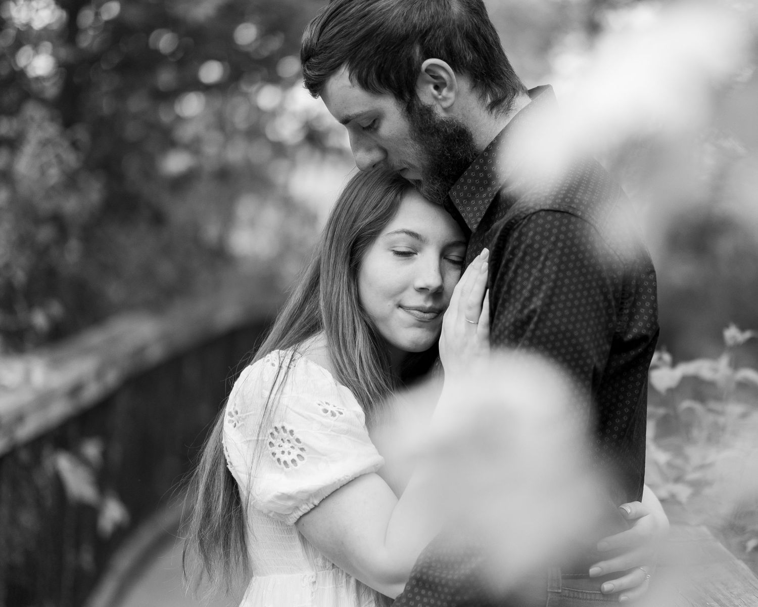 Black and white photo of a couple embracing outdoors, the woman with closed eyes resting her head on the man's chest near a railing.