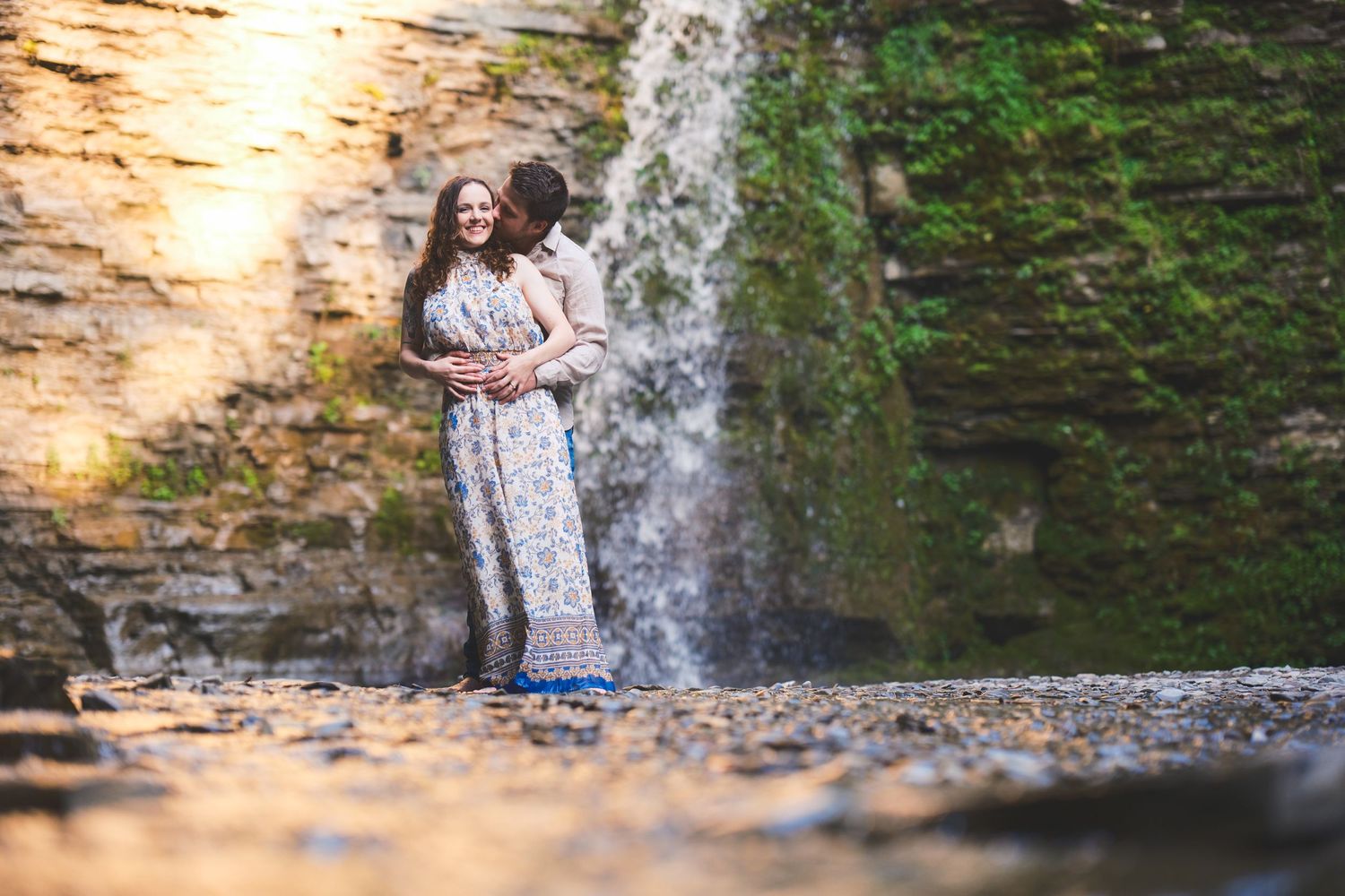 Couple embraces and kisses on a rocky creek bed with a waterfall and mossy cliff behind them in golden light.