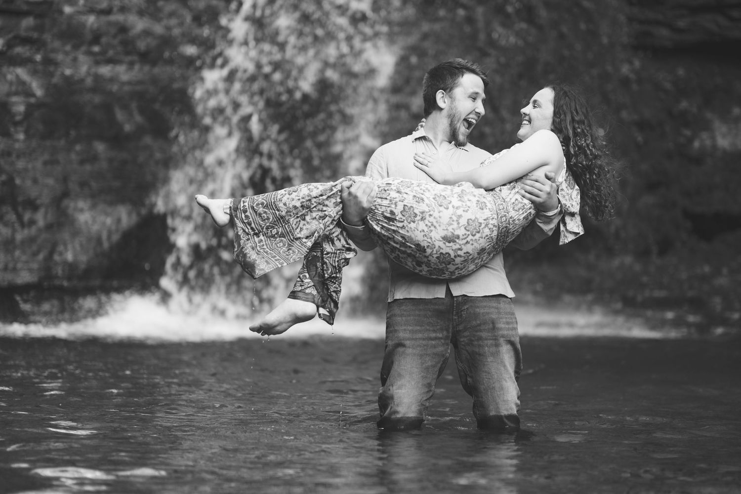 Smiling man standing in shallow water carries a laughing woman in his arms in front of a waterfall in a black and white photo.