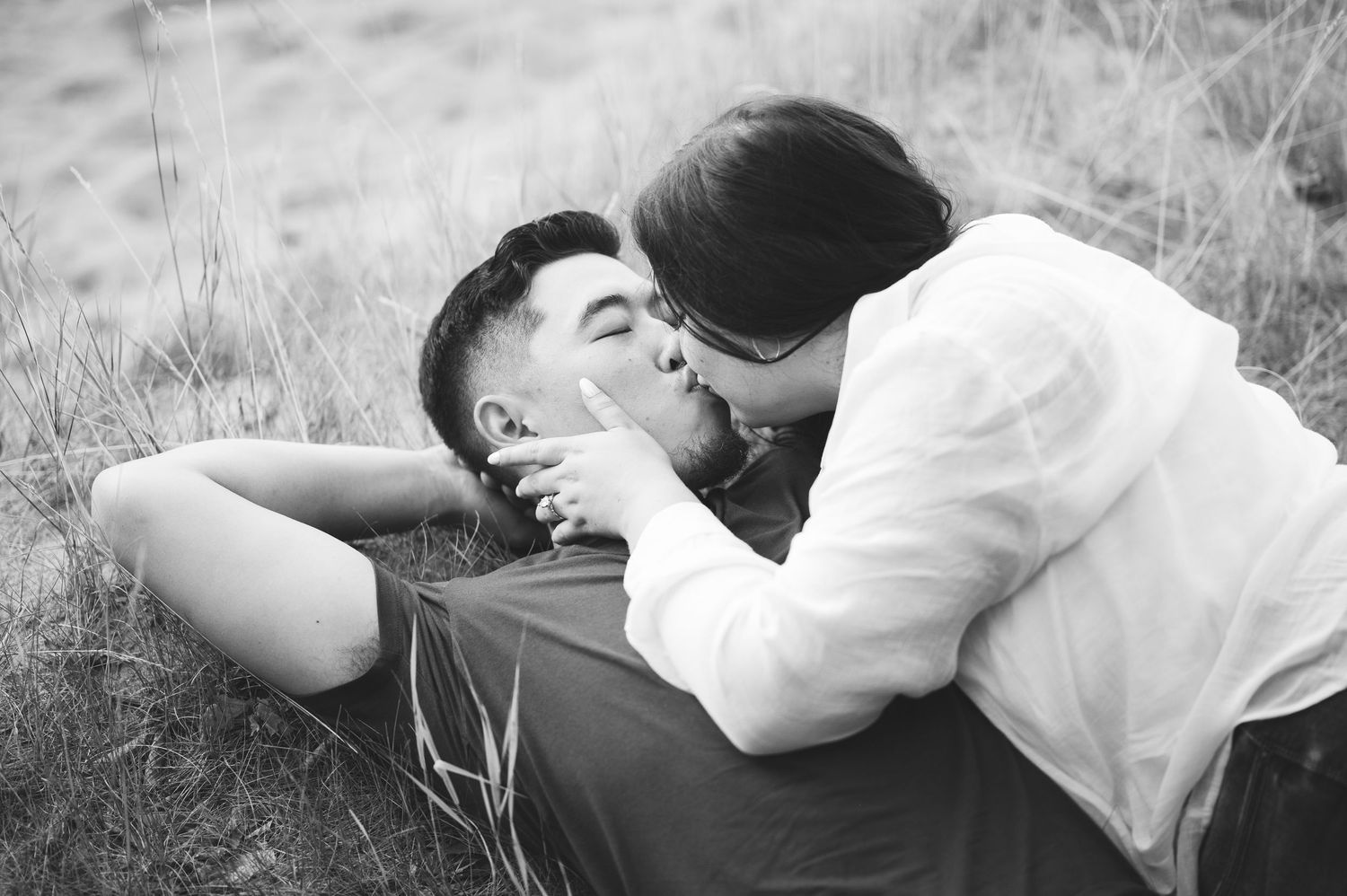 Black and white photo of a couple kissing outdoors as a woman leans over a man lying in tall grass.
