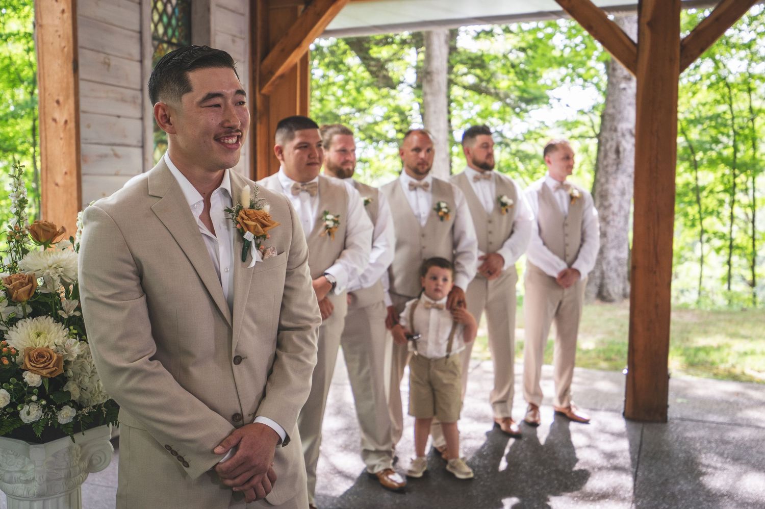 Groom in a tan suit stands smiling in a rustic wooden pavilion with groomsmen and a young ring bearer lined up behind him.