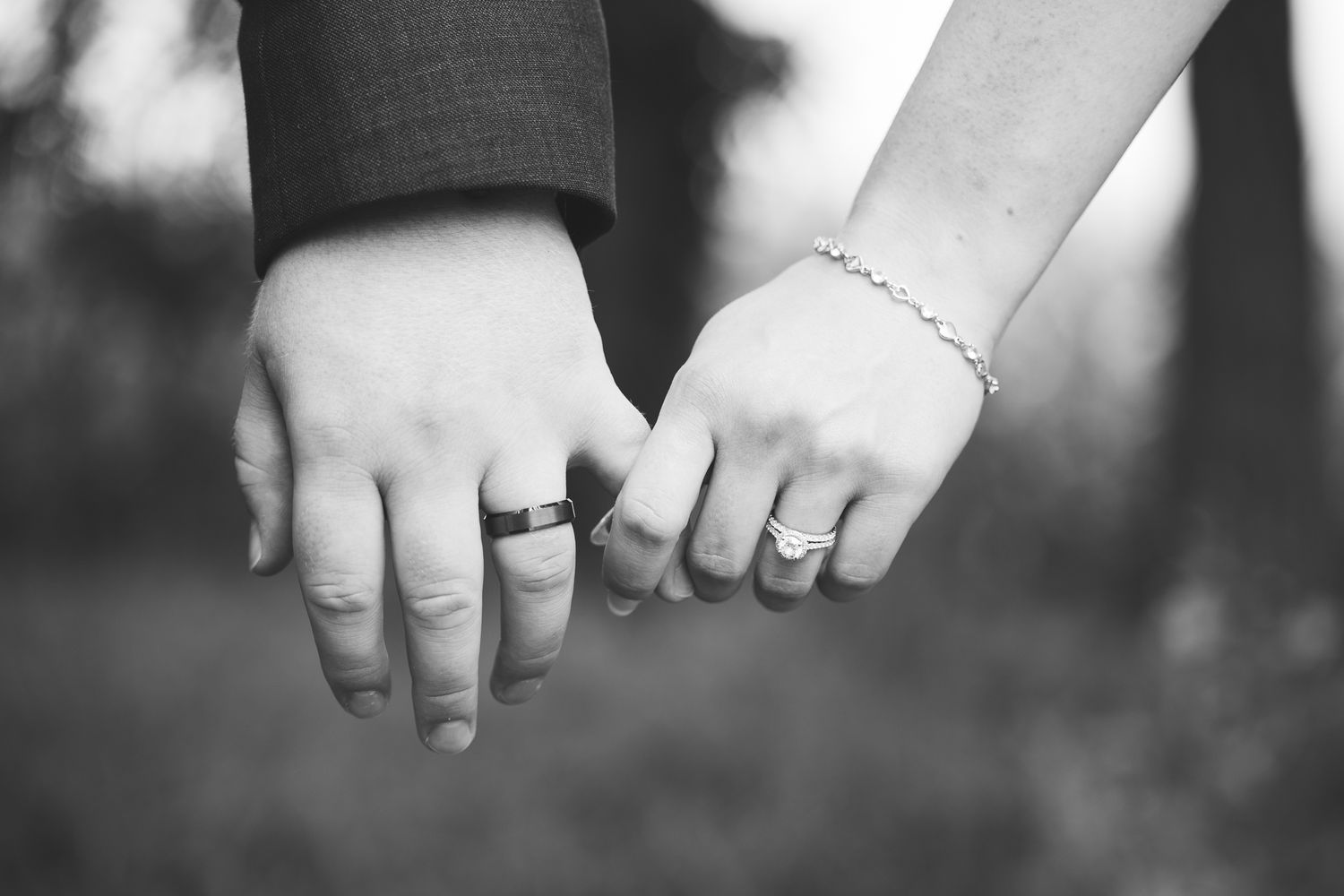 Black and white close-up of a bride and groom holding hands, showing their wedding rings and a delicate bracelet outdoors.