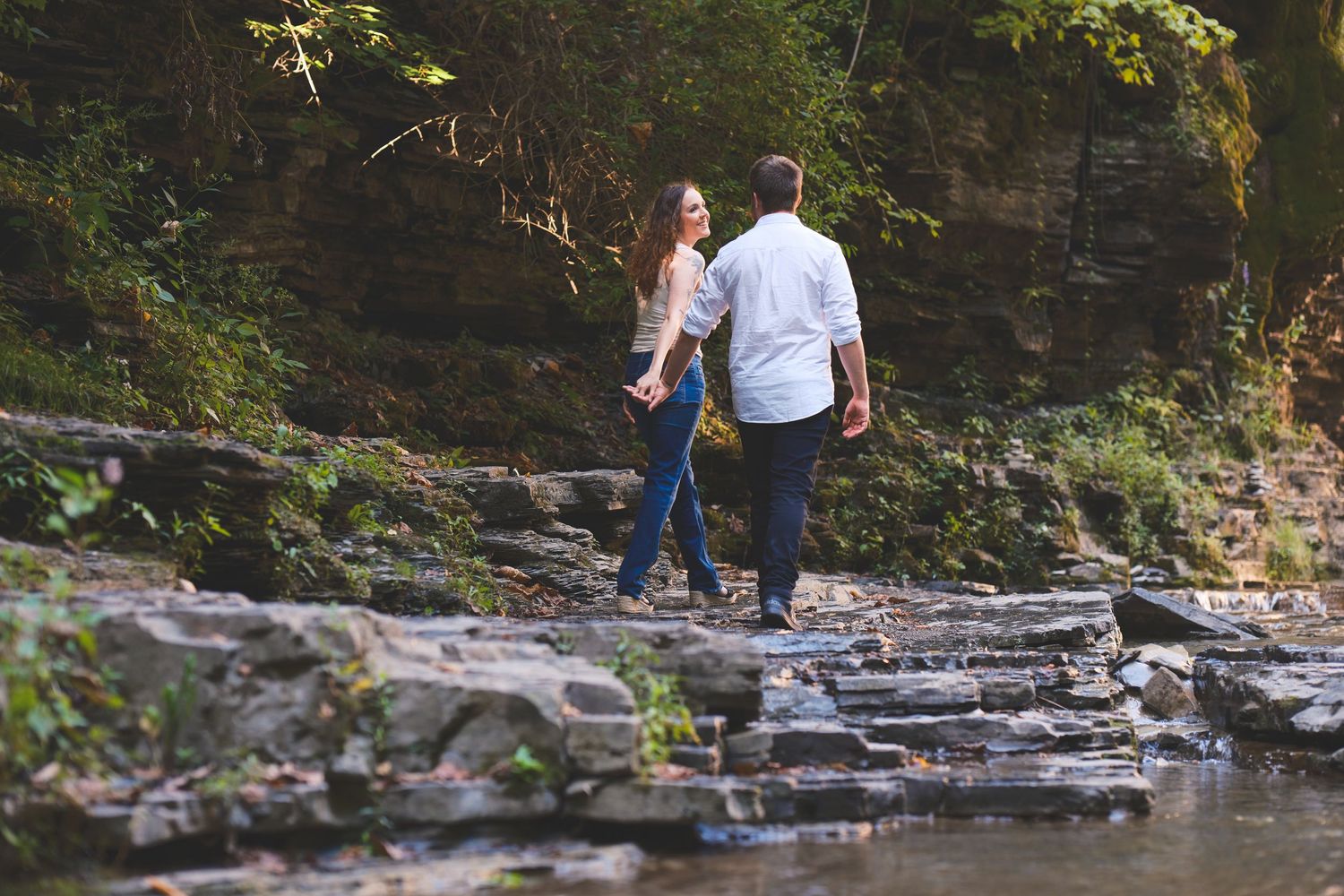 A couple holds hands and walks across flat rocks beside a creek in a shaded wooded gorge during an engagement photo session.
