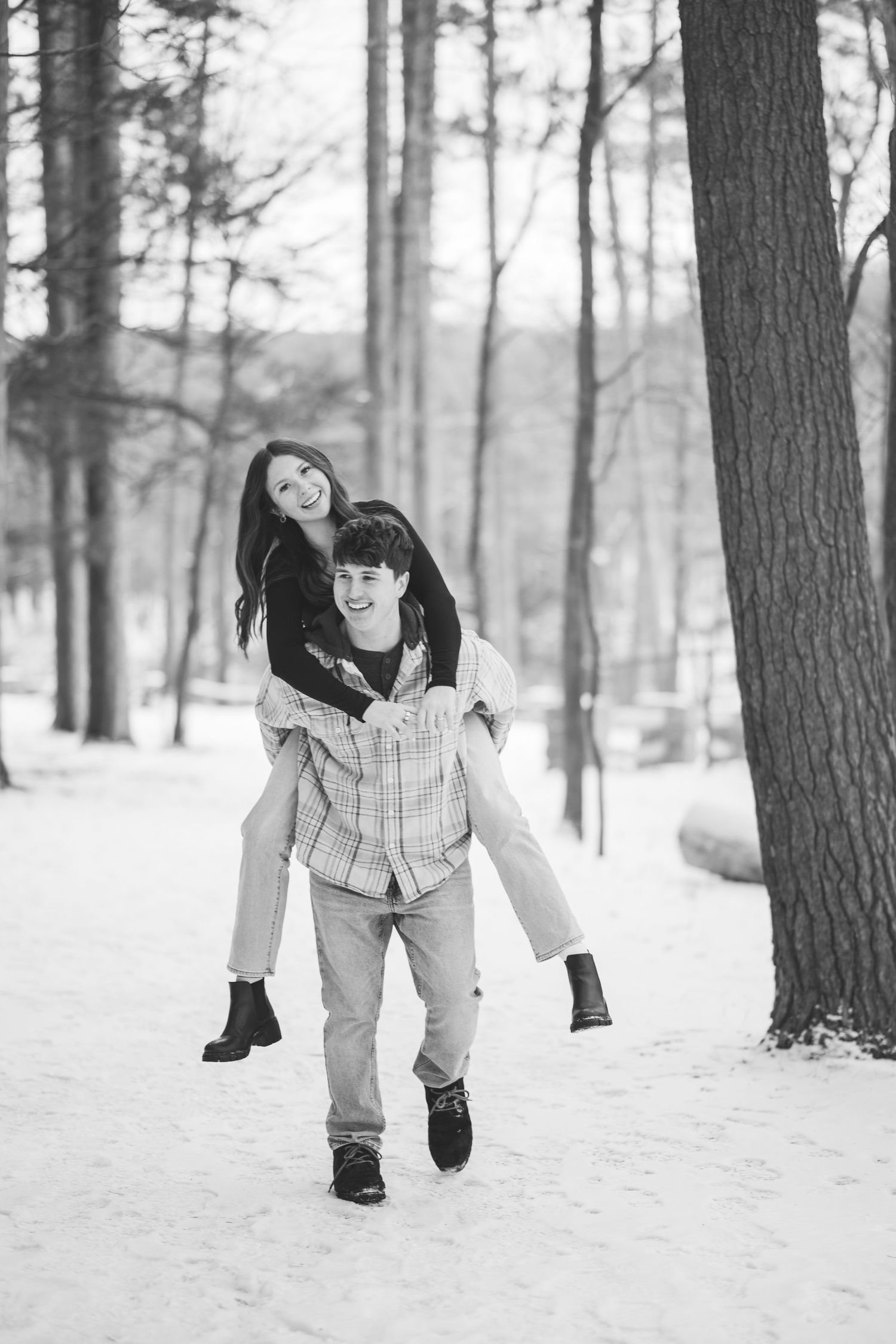 A smiling man gives a laughing woman a piggyback ride through a snowy forest in this black and white engagement photo.