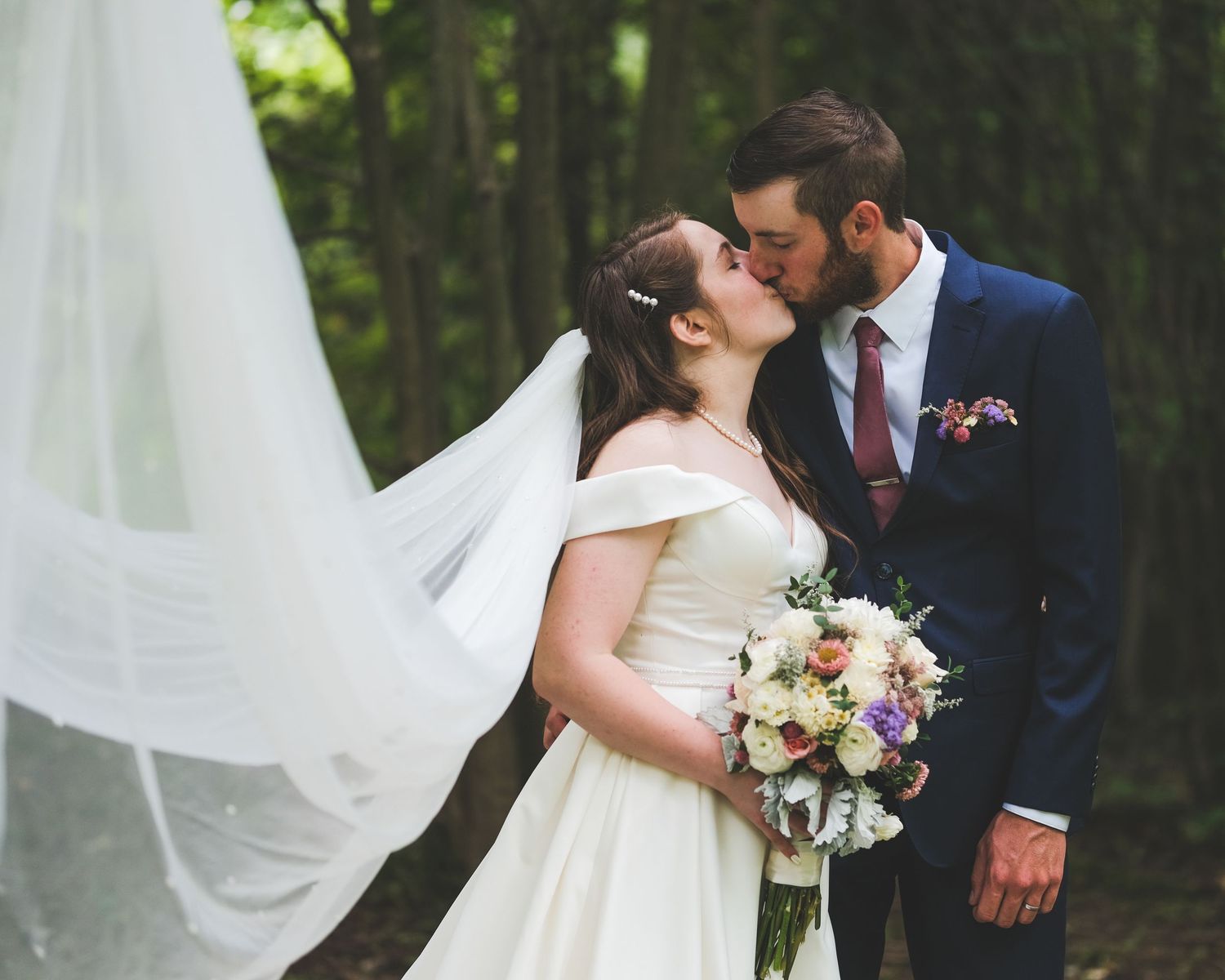 A bride in a white gown and a groom in a navy suit share a kiss in a forest as her long veil blows in the breeze.