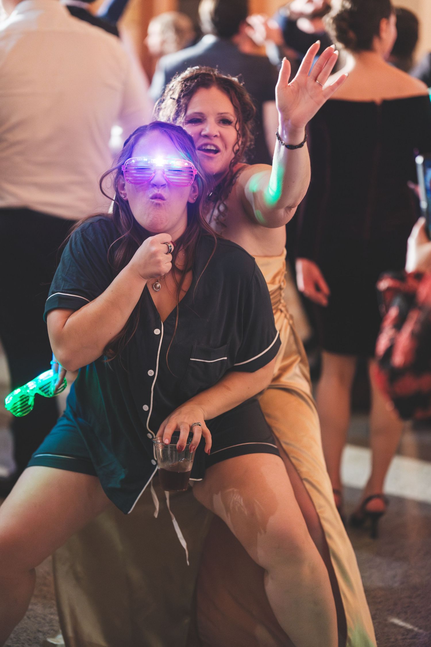 Two women dance and laugh at a lively party, one in glowing pink glasses and navy pajamas holding a drink, the other in a gold gown.