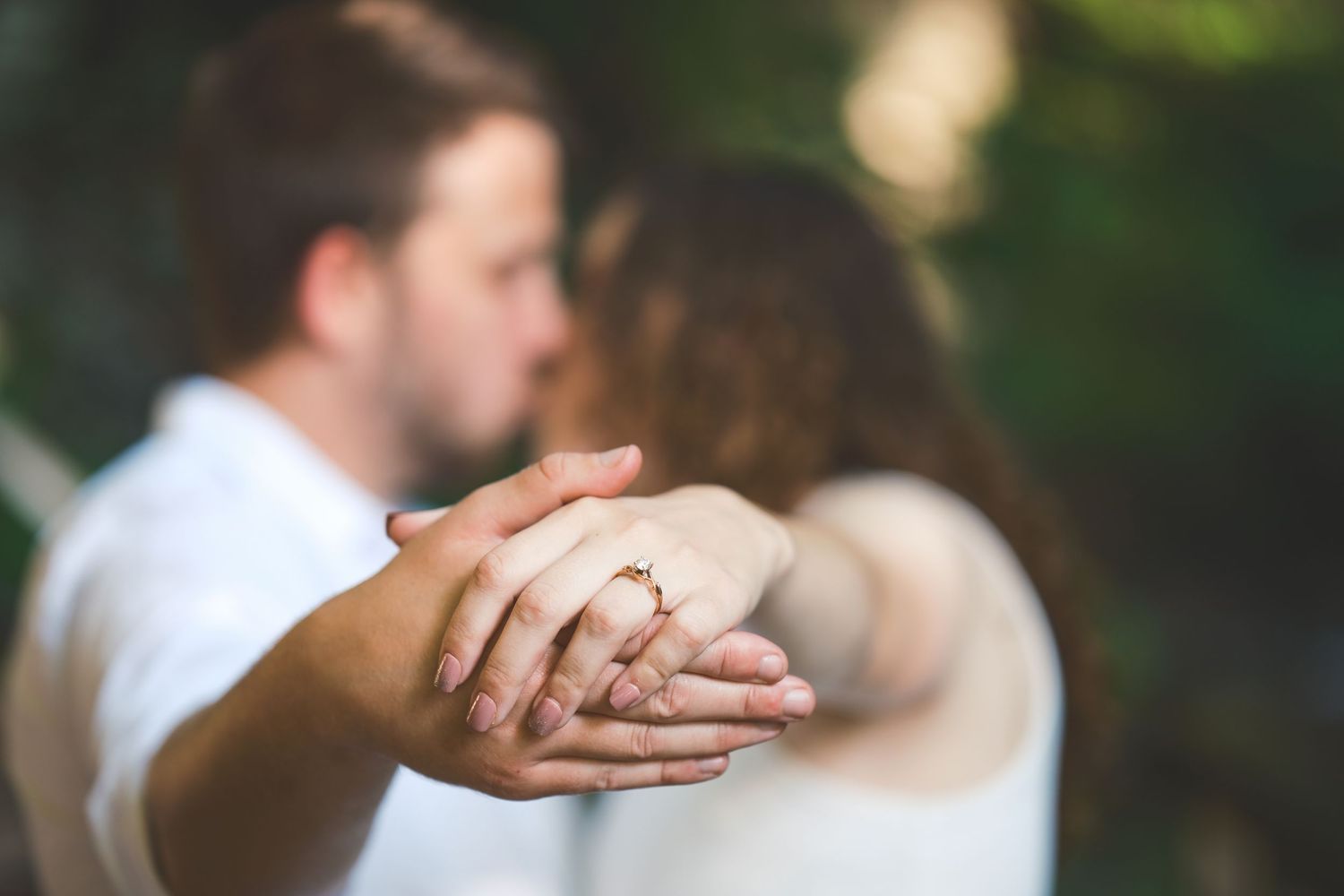 Close-up of a couple holding hands to show an engagement ring while embracing, with soft blurred greenery in the background.