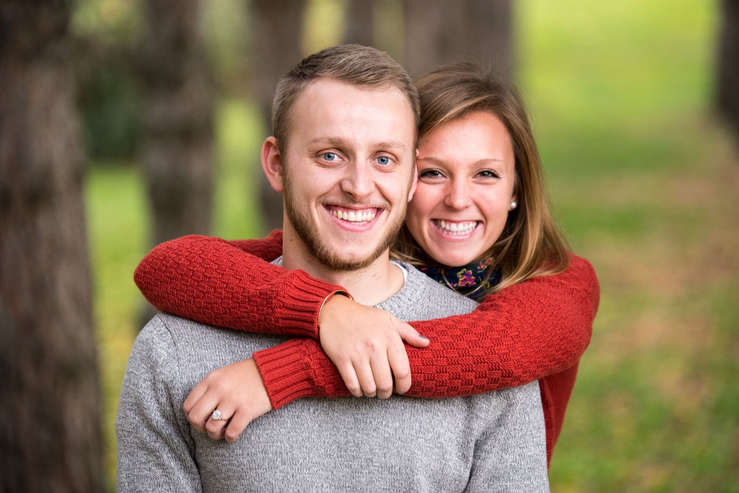Smiling young woman in a red sweater hugs a smiling bearded man in a gray sweater from behind outdoors among trees.