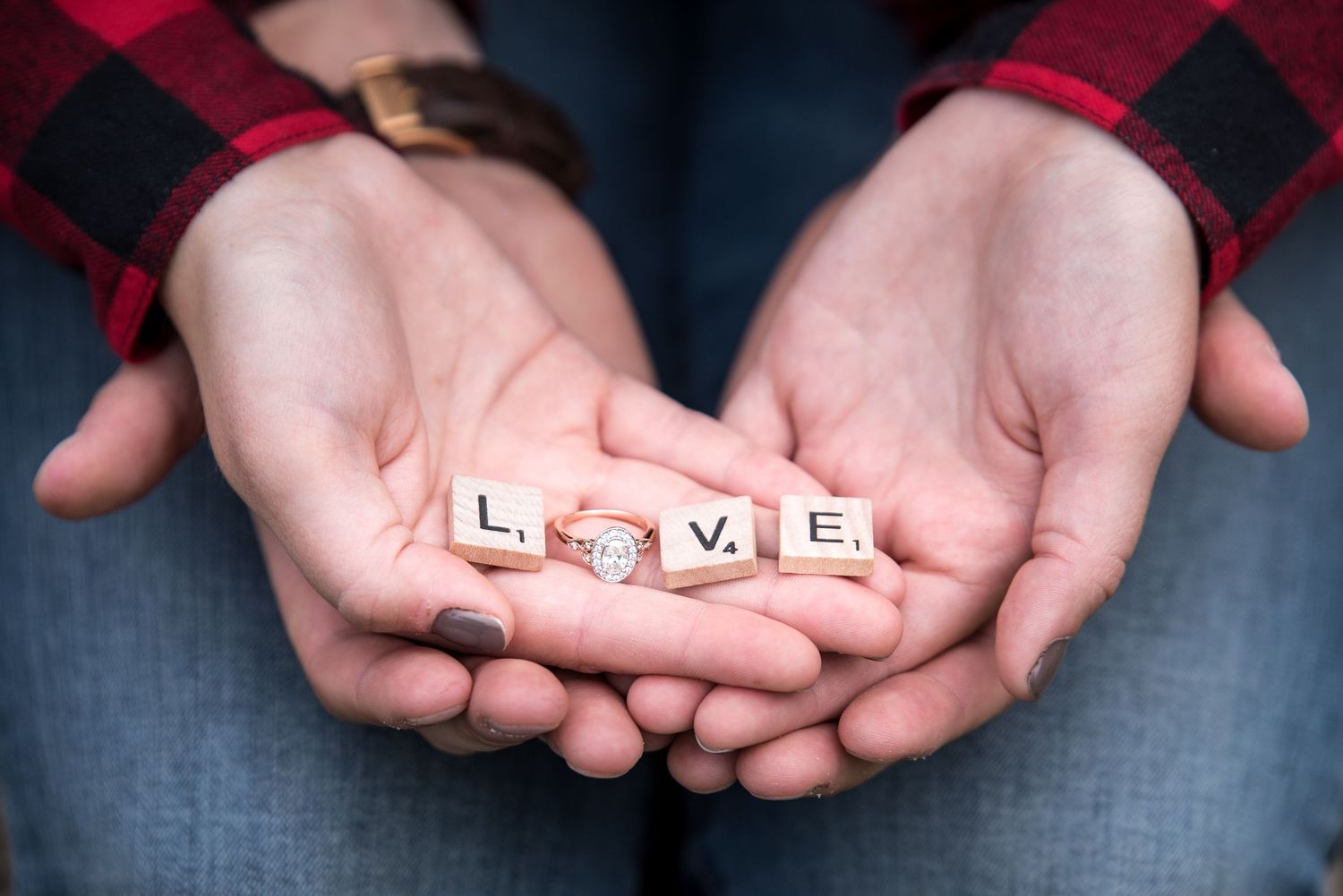 Open hands hold Scrabble tiles spelling LOVE with a diamond engagement ring replacing the O, worn with a plaid flannel shirt.