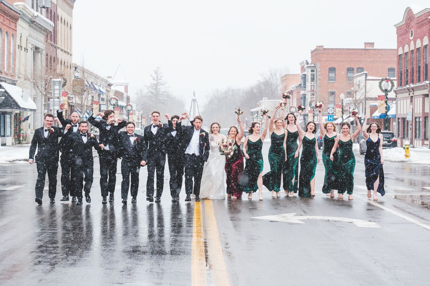 A wedding party in black tuxedos and green dresses walks down a wet city street with the bride and groom, arms raised in celebration.