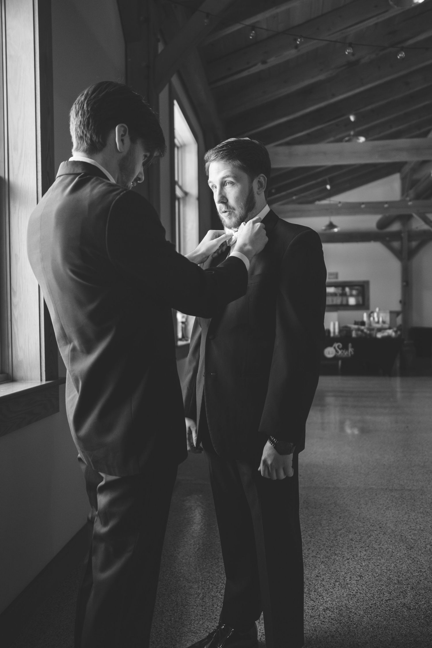 Black and white photo of a groomsman adjusting a groom's bow tie in a rustic venue with wood beams and large windows.