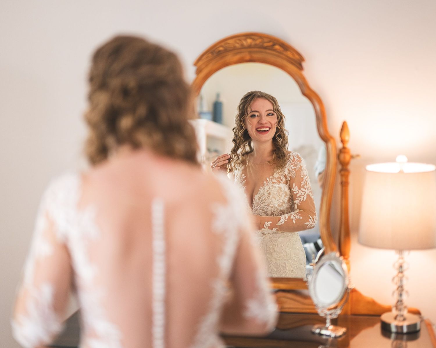 Bride in a lace wedding gown smiles at her reflection in an ornate wooden mirror while getting ready for her wedding.