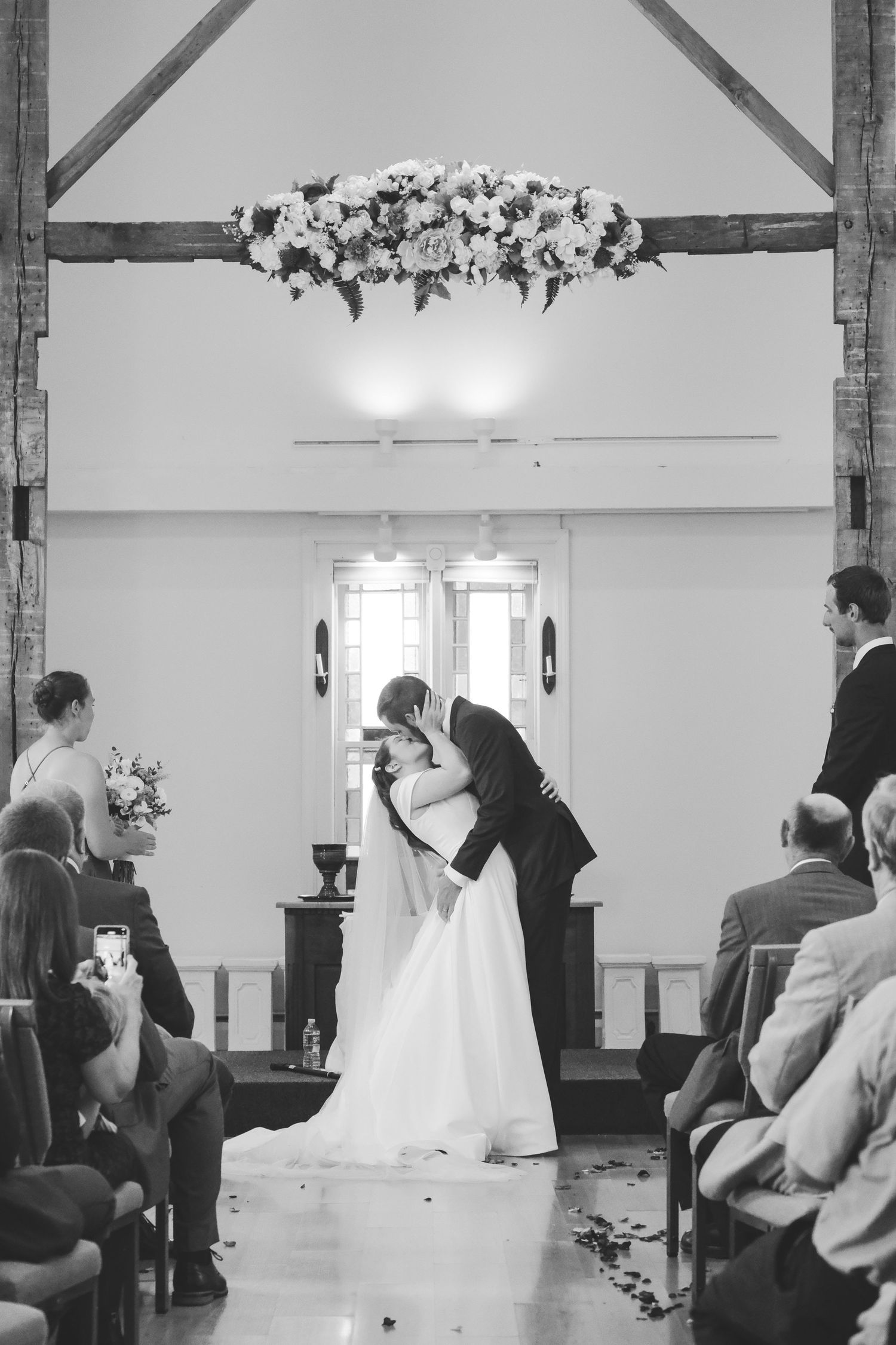 Black and white photo of a bride and groom sharing their first kiss at the altar beneath a floral arrangement as guests watch.