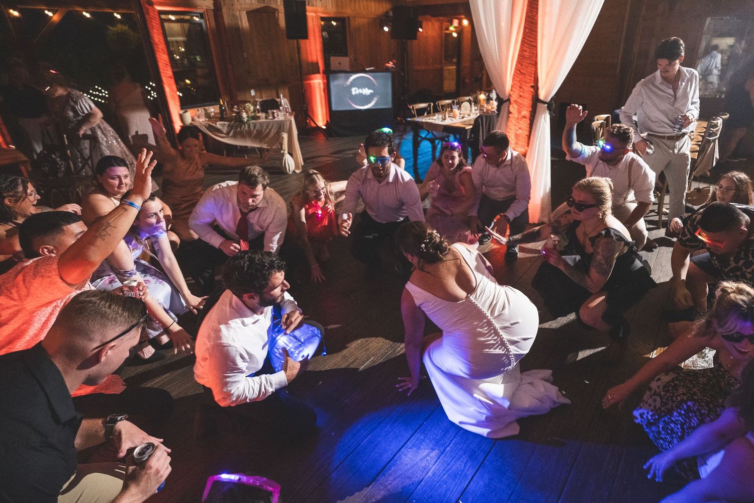 Wedding guests sit in a circle on a dim dance floor cheering as a bride in a white gown dances in the center.