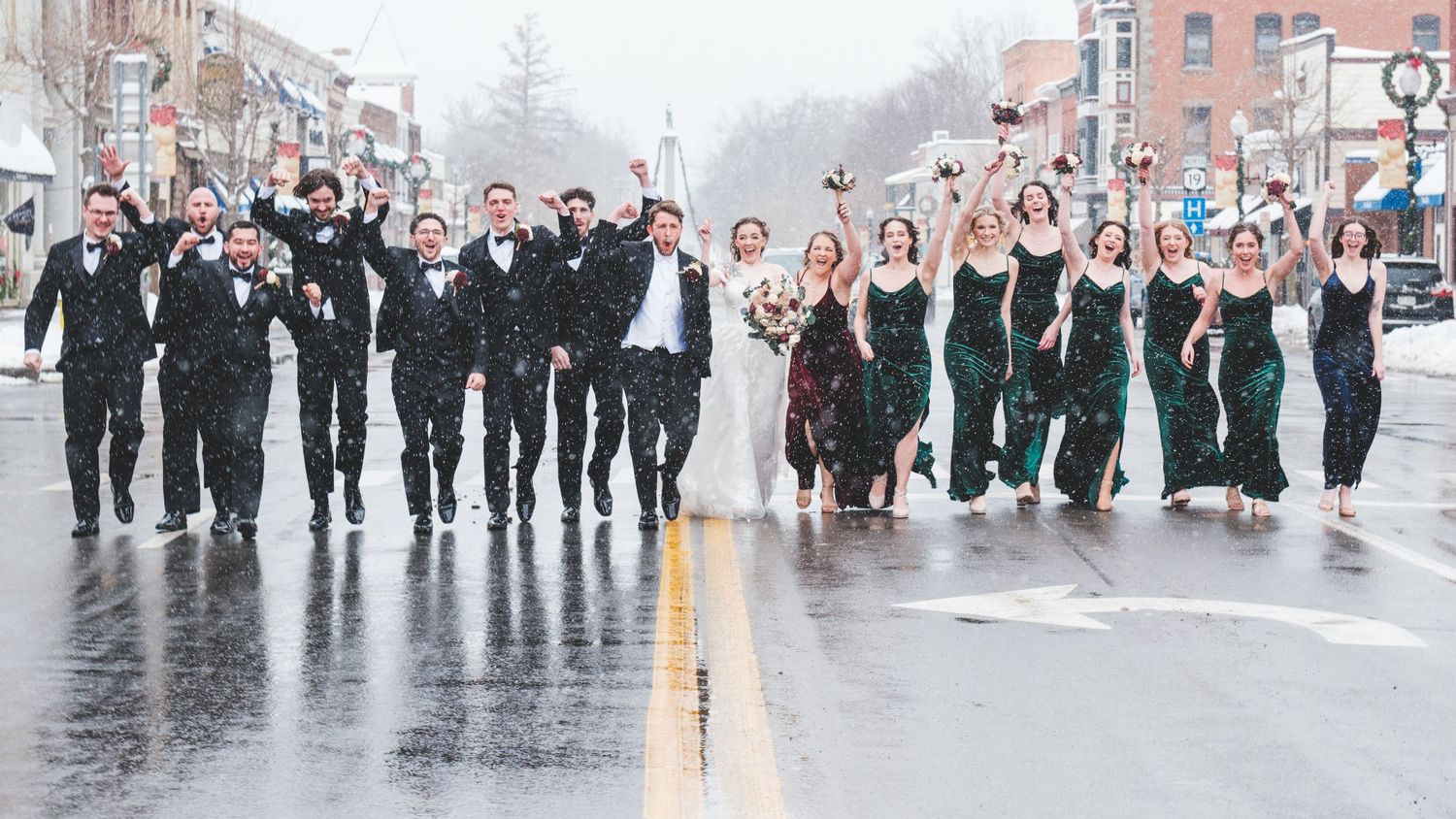 Wedding party in black tuxedos and emerald green dresses walks with the bride and groom down a rainy city street.
