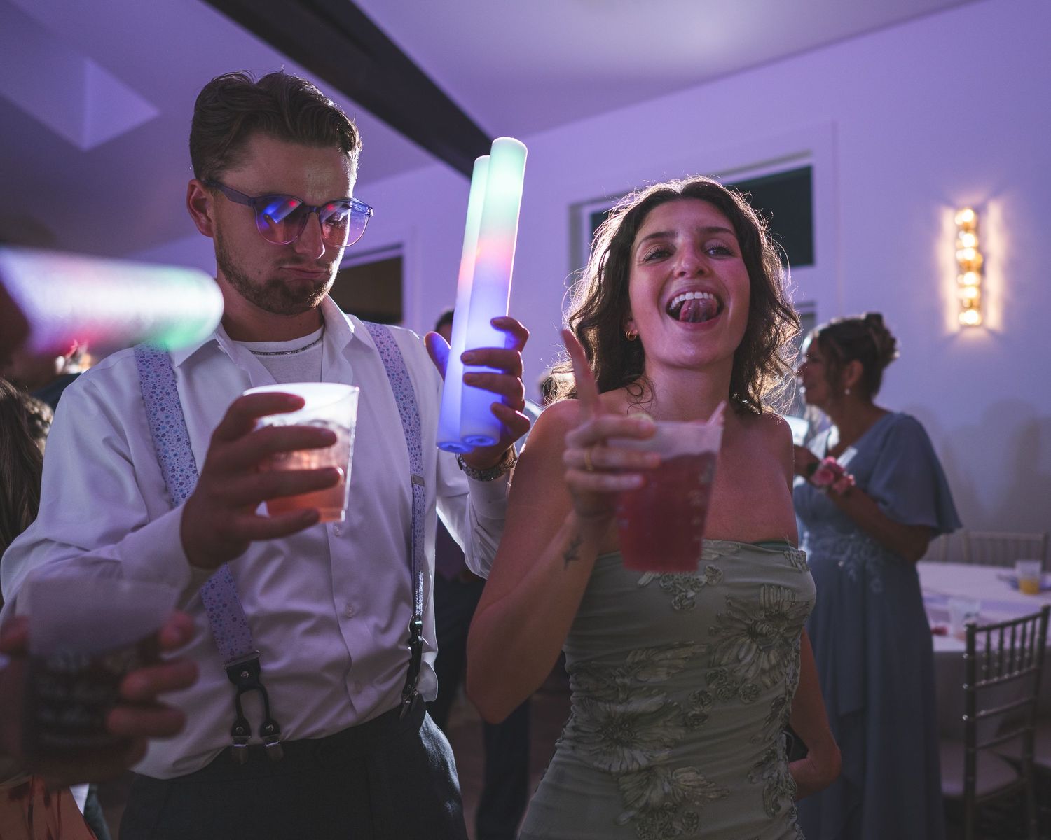 A man with glasses holding a glow stick and a woman laughing with a drink dance together at a lively wedding reception.