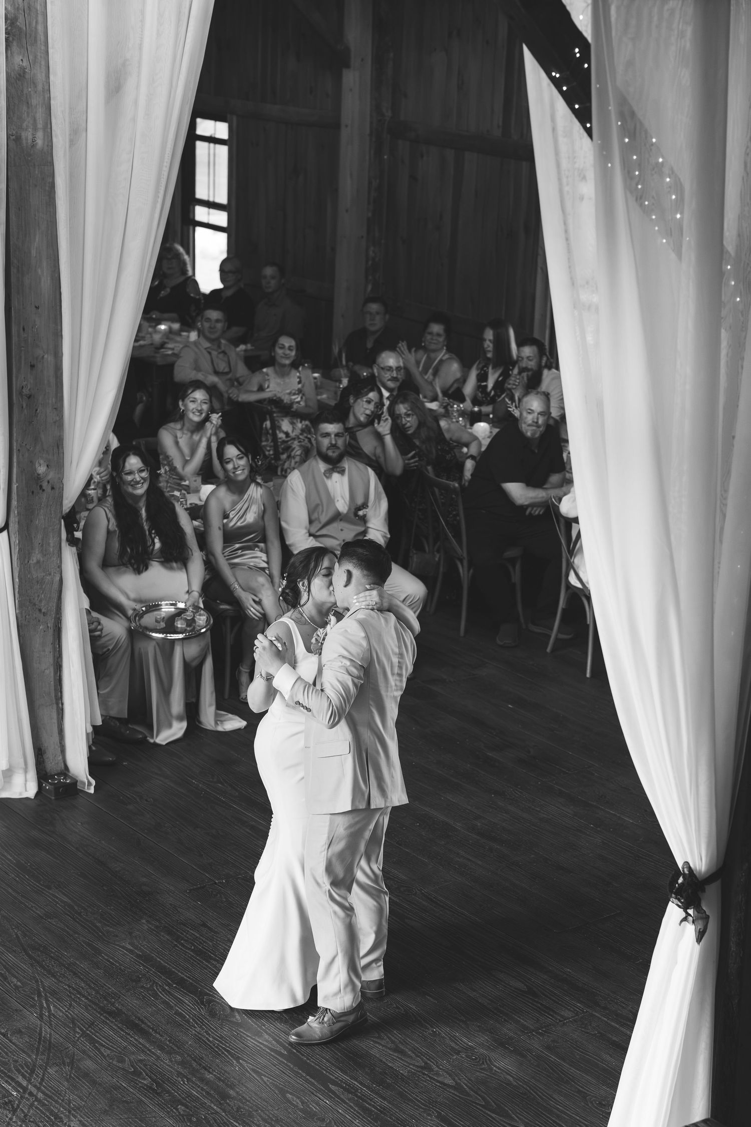 A black and white photo of a couple sharing a first dance in a rustic barn while seated guests watch from behind white drapes.