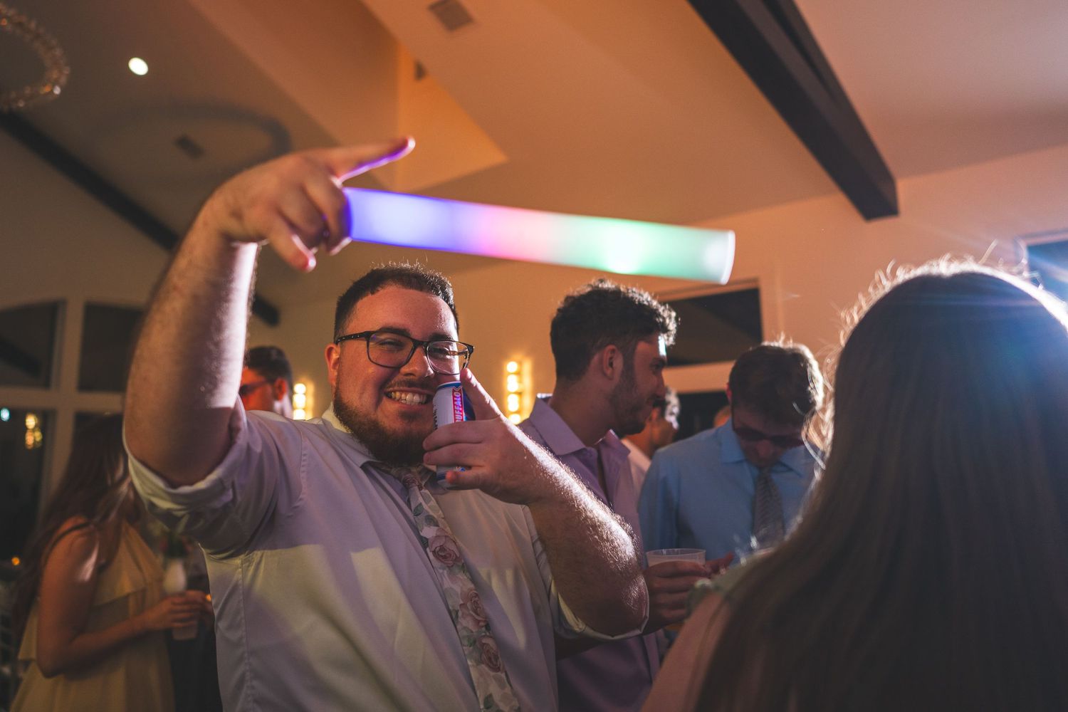 Smiling man with glasses points and holds a glowing light stick while dancing with guests at a wedding reception.
