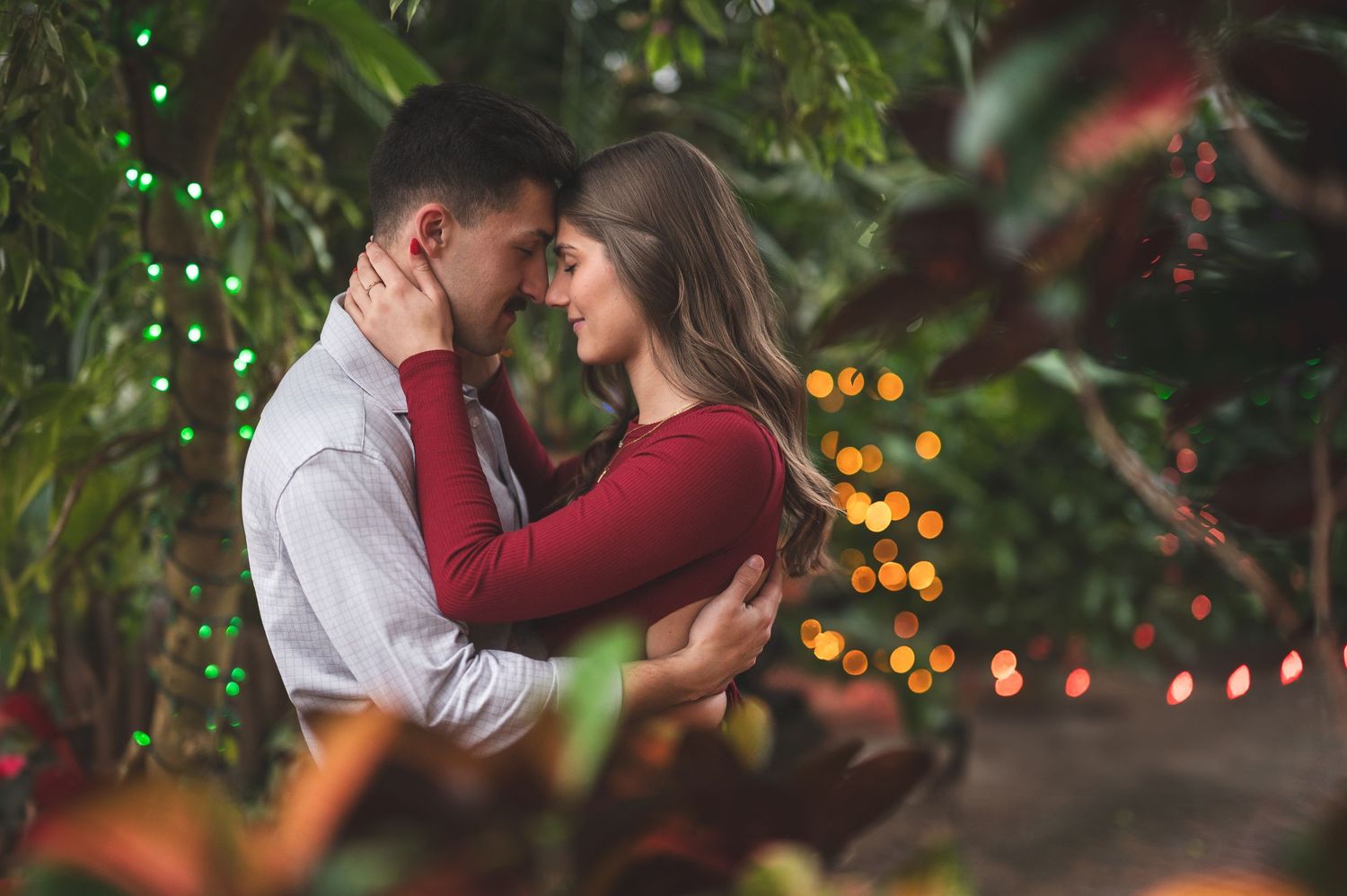 Engaged couple embracing and nearly kissing among lush greenery and warm glowing string lights during an outdoor engagement session.