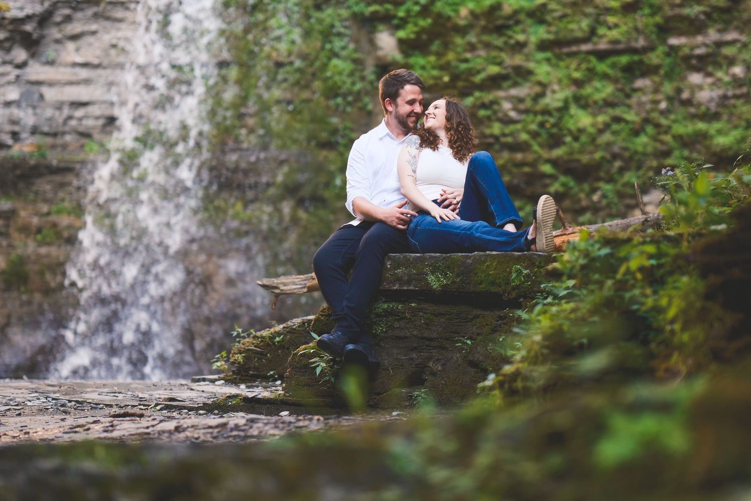 Smiling couple sits close together on a mossy stone ledge beside a cascading waterfall surrounded by lush green forest.