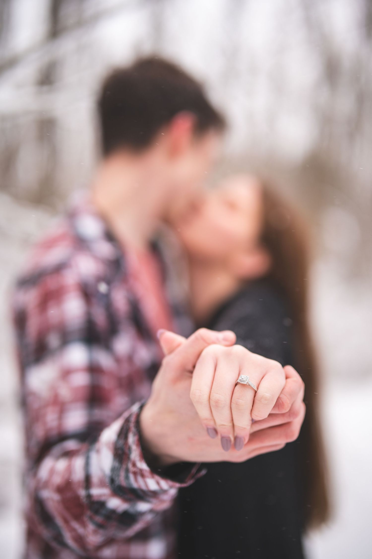 Couple kissing in a snowy forest while their clasped hands show off a diamond engagement ring in sharp focus.