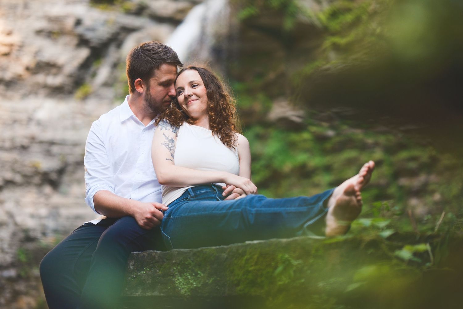 A couple sits close together on a mossy rock near a waterfall, smiling and embracing during an outdoor engagement photo session.