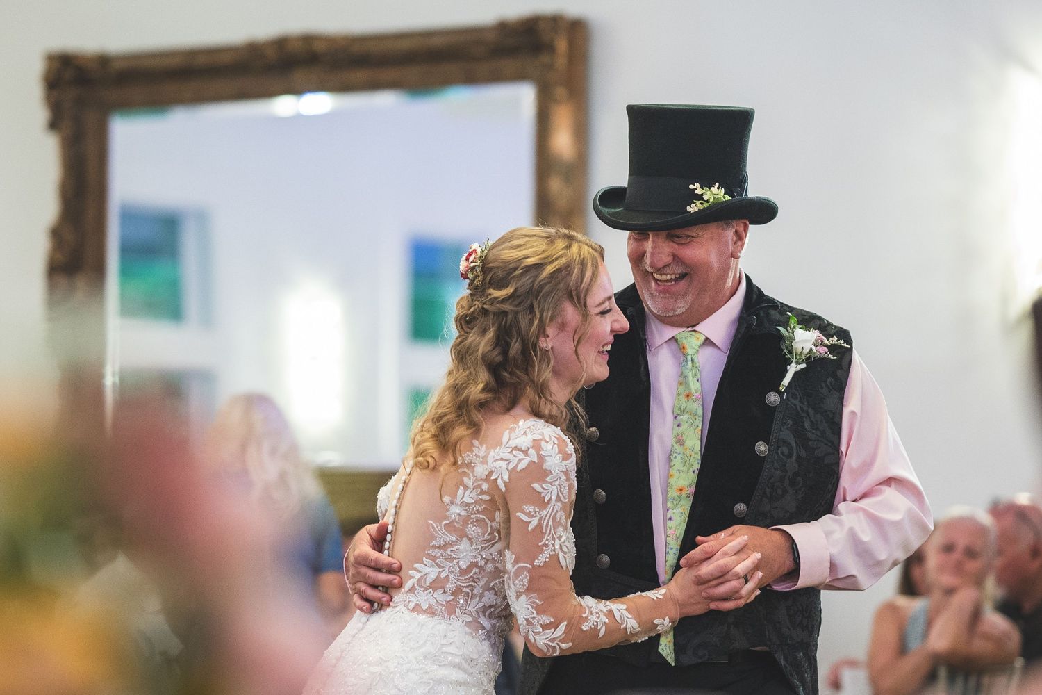 Bride in a lace gown dances with a smiling man in a top hat and vest during a wedding reception dance.