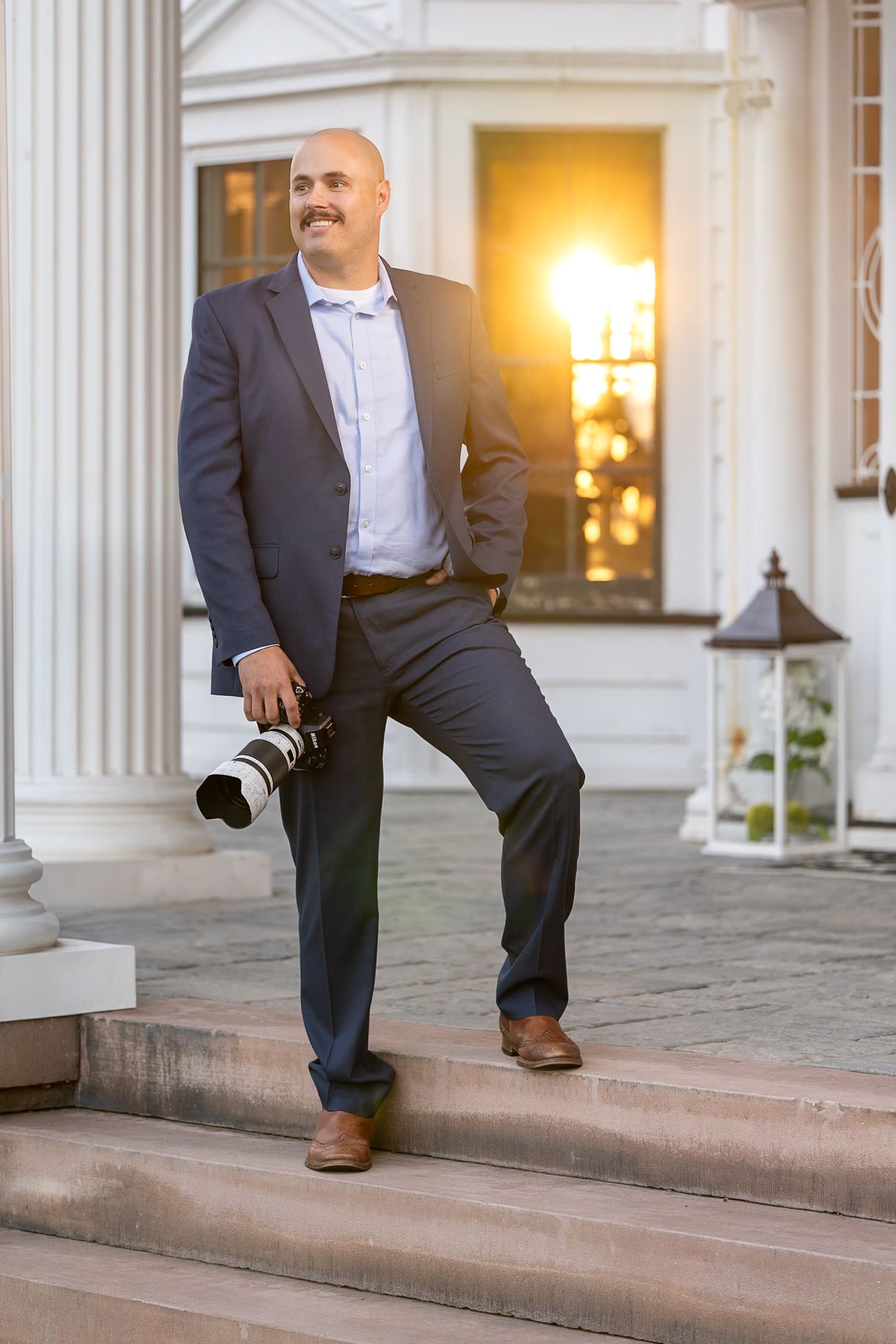 Smiling bald man in a navy blazer and brown shoes holds a camera with a telephoto lens while standing on stone steps near white columns.