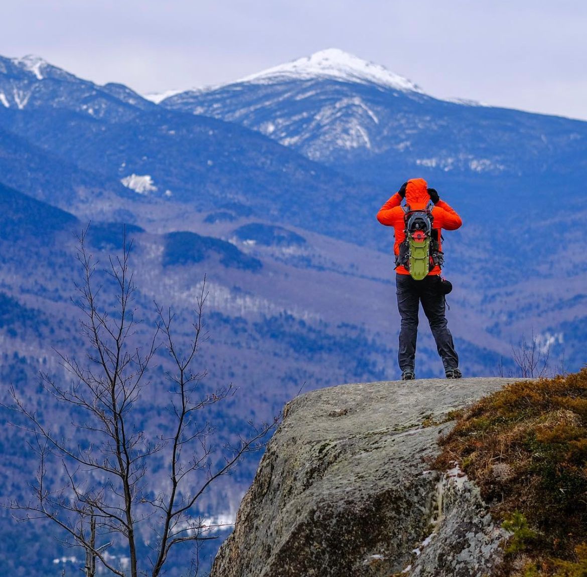 Hiker in an orange jacket and green backpack stands on a rocky outcrop viewing snow-dusted mountain peaks in the distance.