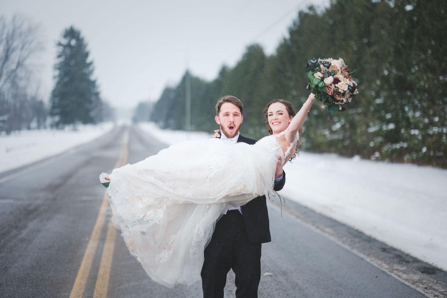 A groom carries his bride in a white wedding dress down a snowy road as she raises her floral bouquet in the air.